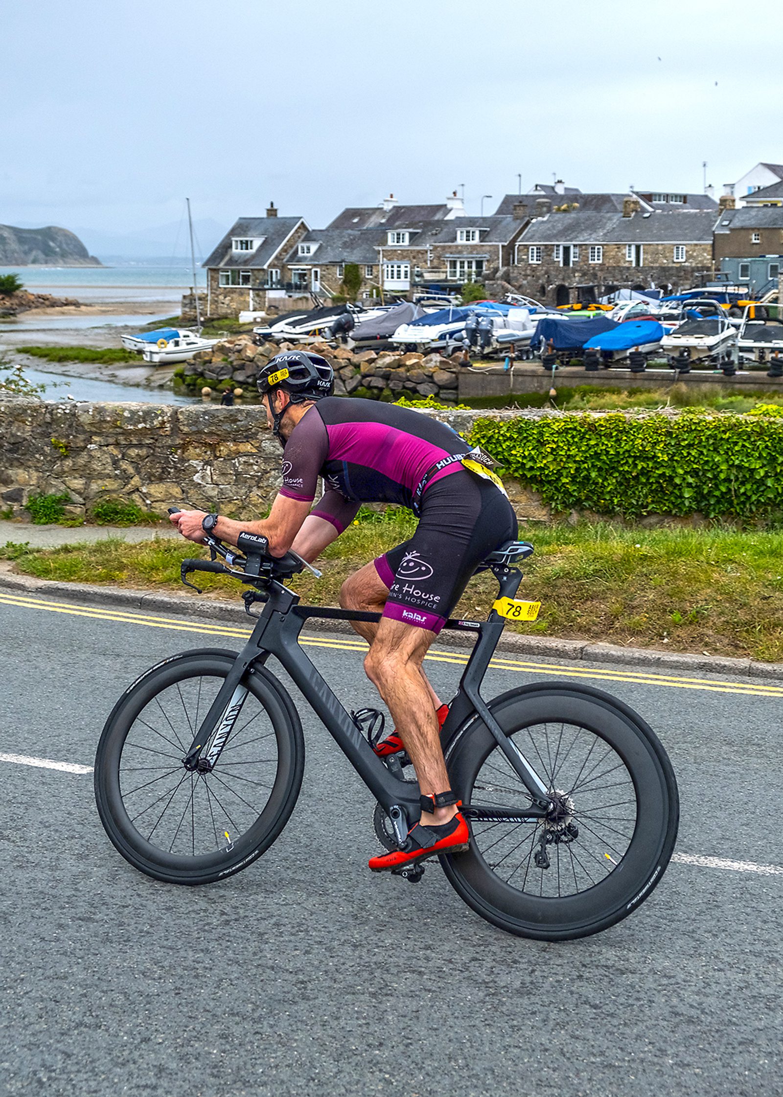 A cyclist wearing a black helmet and a purple and black cycling outfit rides a black bicycle on a paved road. The background features a harbor with boats, stone houses, and a grassy area near the road. The sky is overcast.