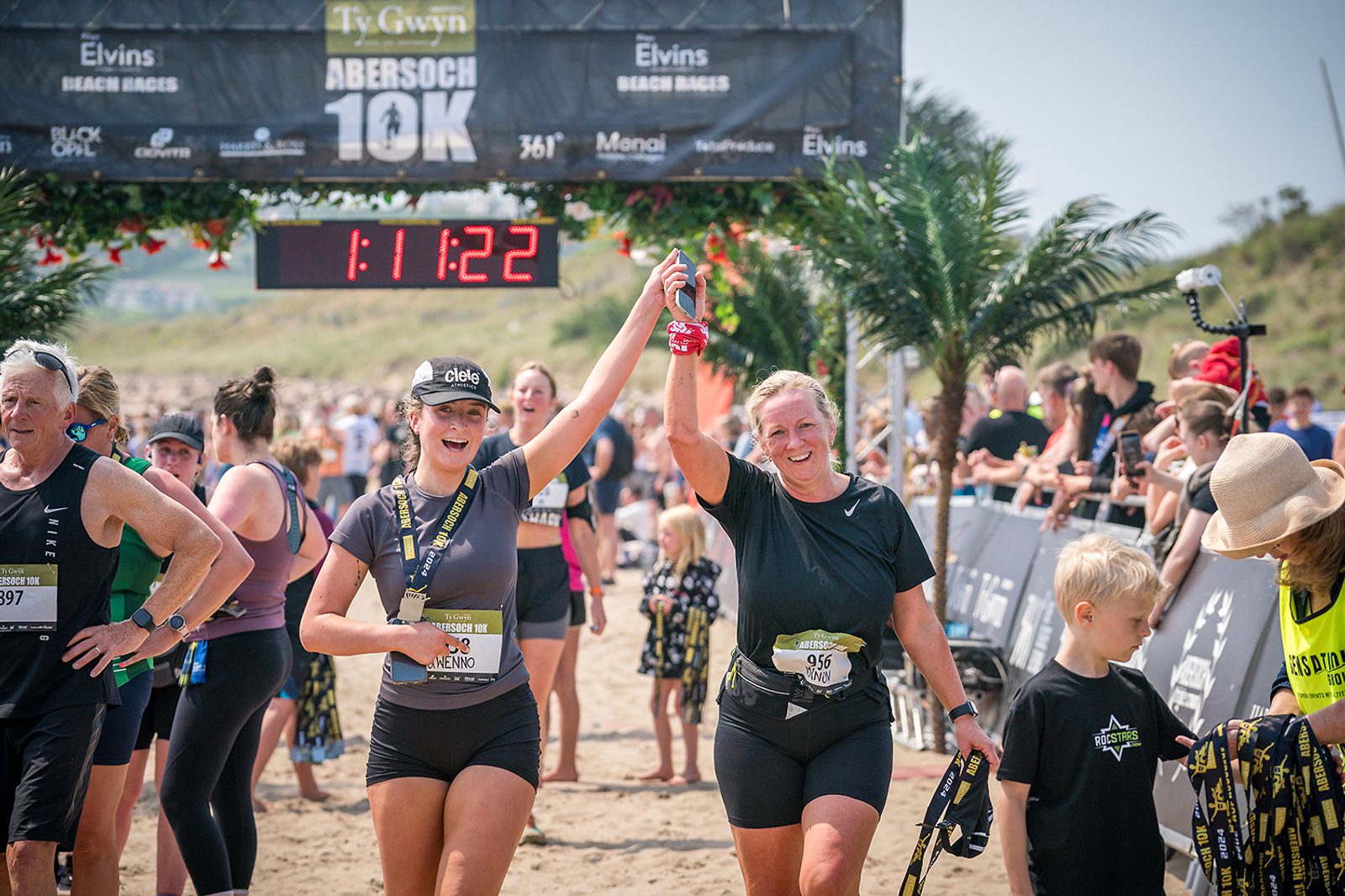 Two women celebrate as they cross the finish line of the Abersoch 10K race holding hands. Other runners and spectators are visible in the background. A timer above the finish line displays "1:11:22". The event seems festive, with greenery and banners.