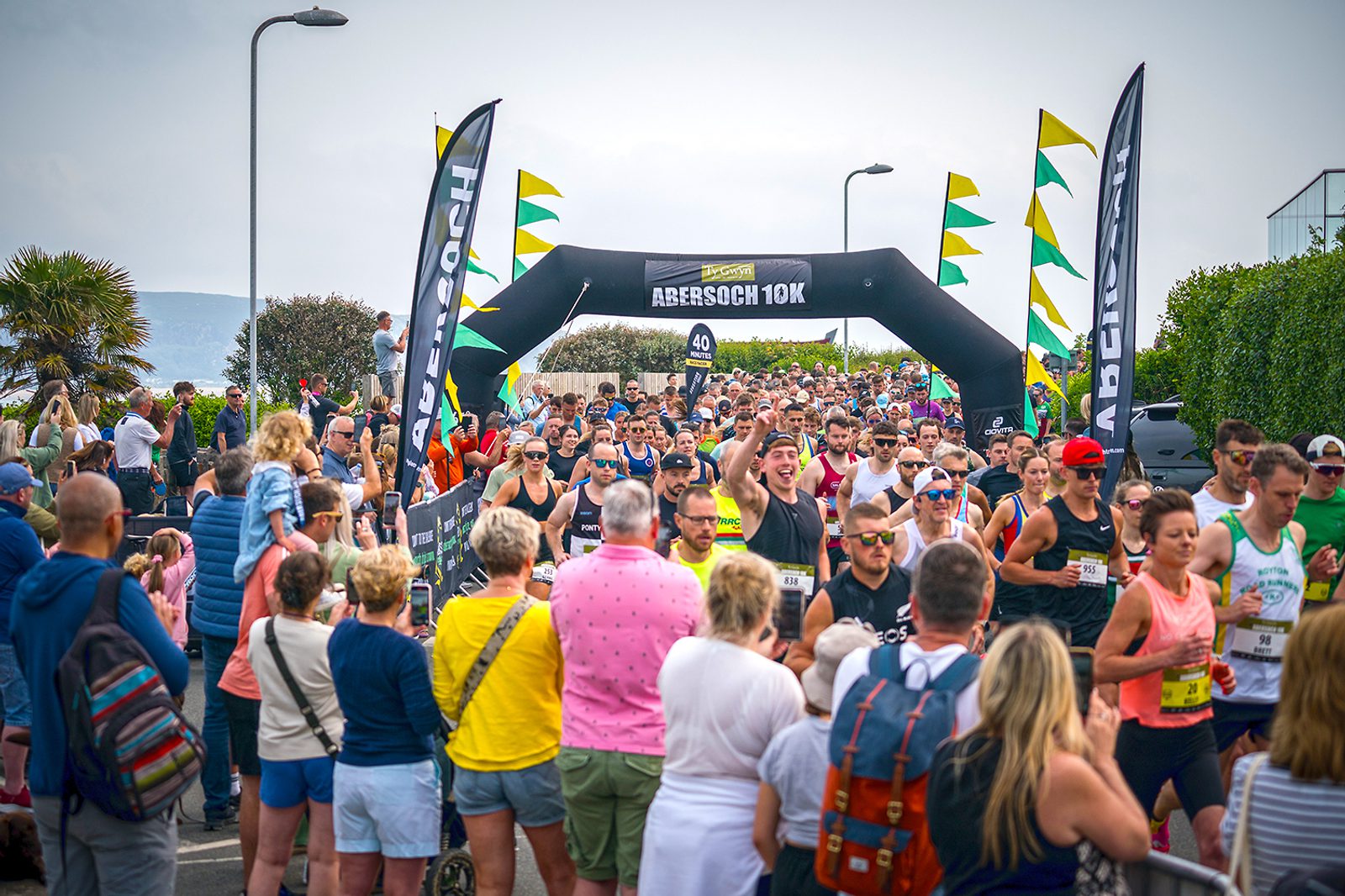 A crowd of runners participate in the Abersoch 10K race, passing through a black inflatable archway with the event's name. Spectators line the sides of the street, cheering and taking photos. The sky is overcast, and the atmosphere is lively and festive.