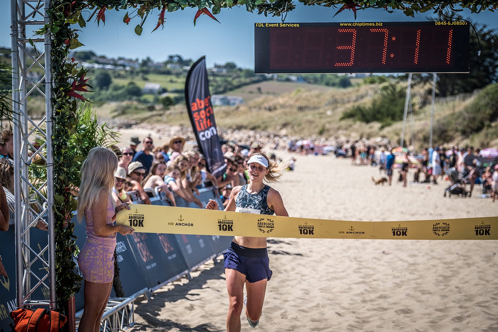 A woman in athletic gear triumphantly crosses the finish line at a beach race, holding a tape with both hands. Spectators cheer behind barriers. A digital timer above her displays "37:11". The background features dunes, flags, and a sunny blue sky.