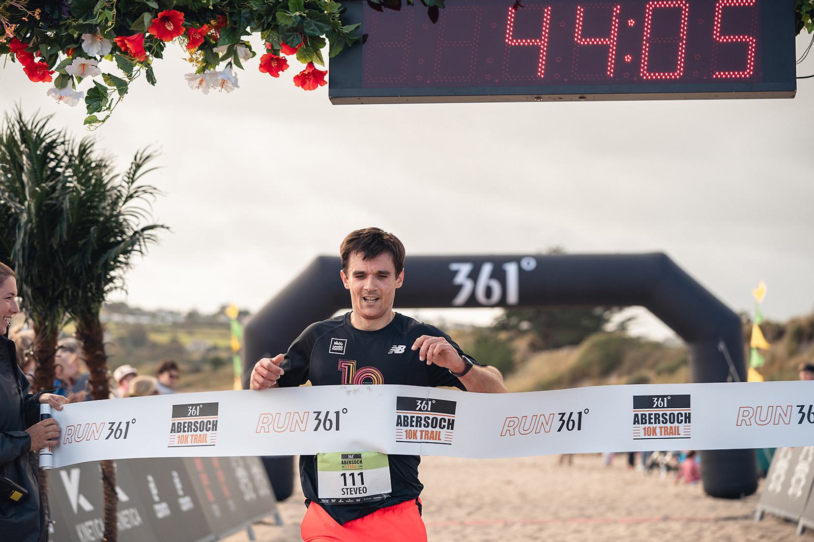 A runner crosses the finish line at the Abersoch 10K Trail, breaking the tape with a digital clock overhead showing 44:05. The background includes spectators, a blow-up archway, and floral decorations.