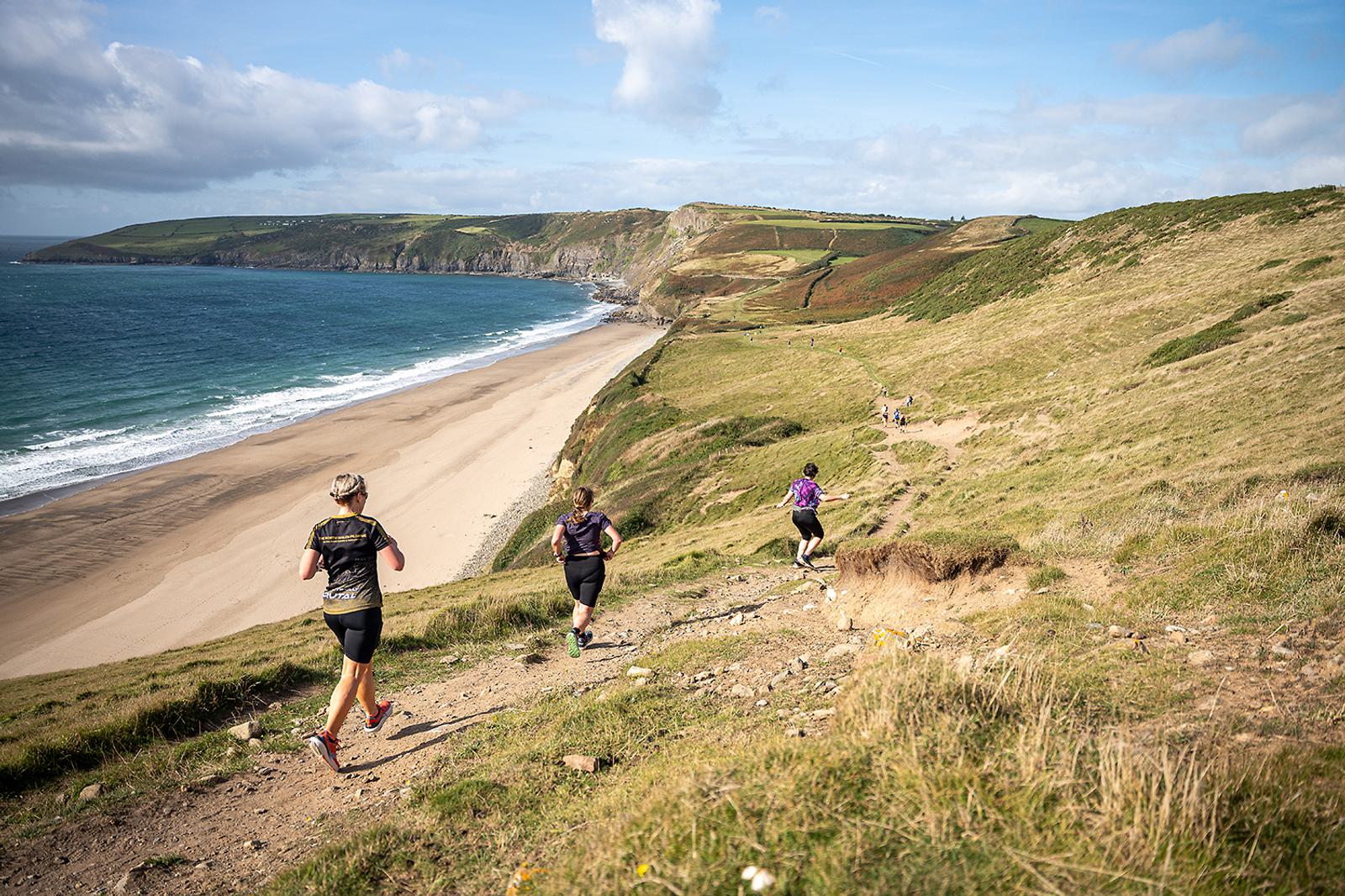 Three people run on a grassy trail overlooking a beach and ocean under a partly cloudy sky. The coastline is lined with cliffs and green hills. Paths and trails can be seen winding through the landscape.