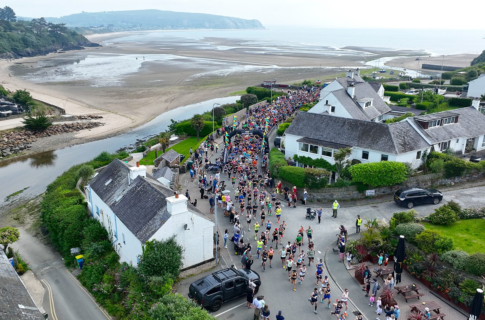 Aerial view of a coastal town during a marathon. Hundreds of runners in colorful attire race along a street lined with white houses surrounded by green hedges. Sandy beaches and the sea are visible in the background, along with distant hills under a cloudy sky.