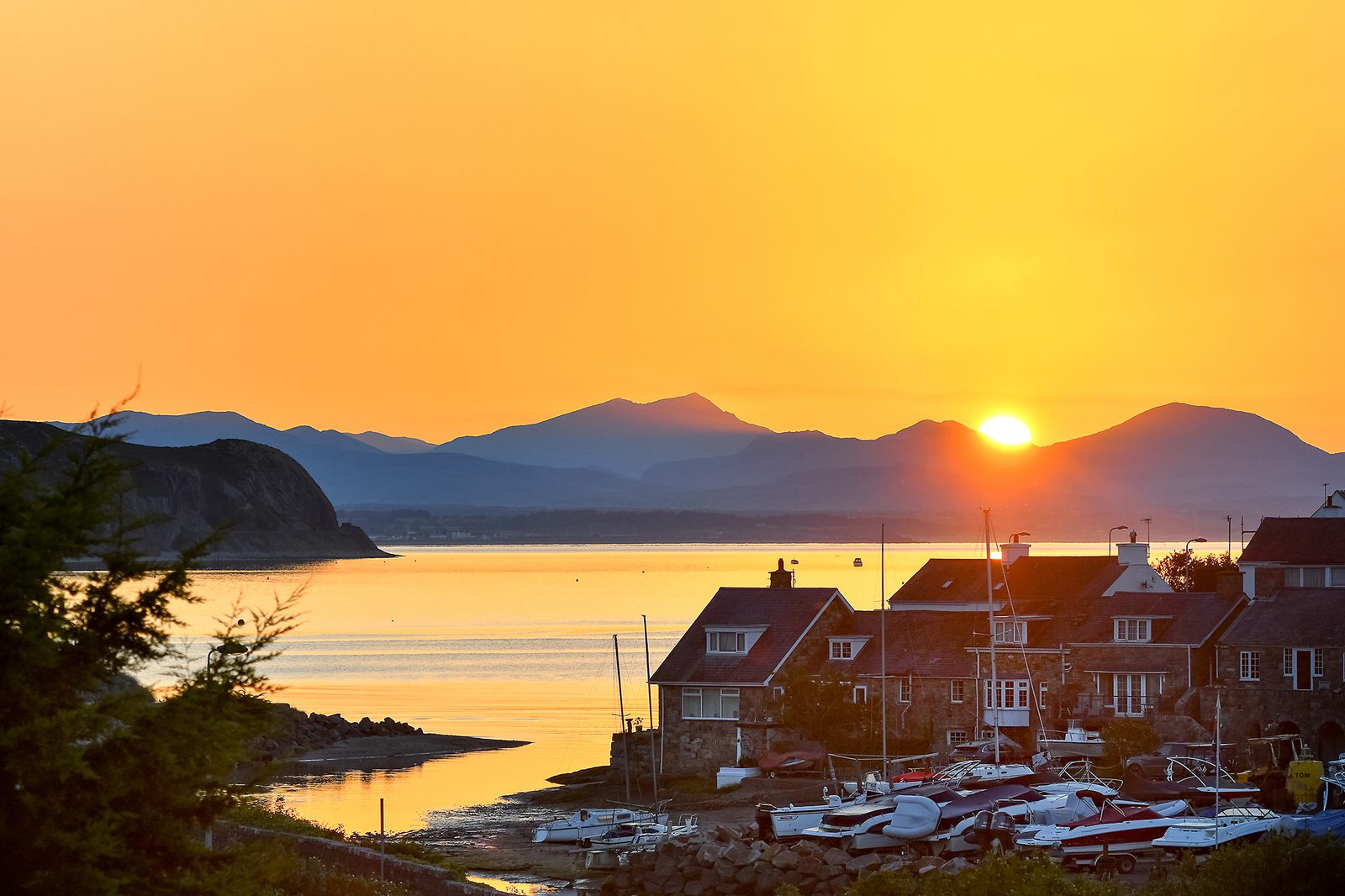 A picturesque sunset over a coastal village, with the sun setting behind distant mountains. The sky is a vibrant orange, casting a warm glow over houses and boats near a calm body of water. The scene is serene and tranquil.