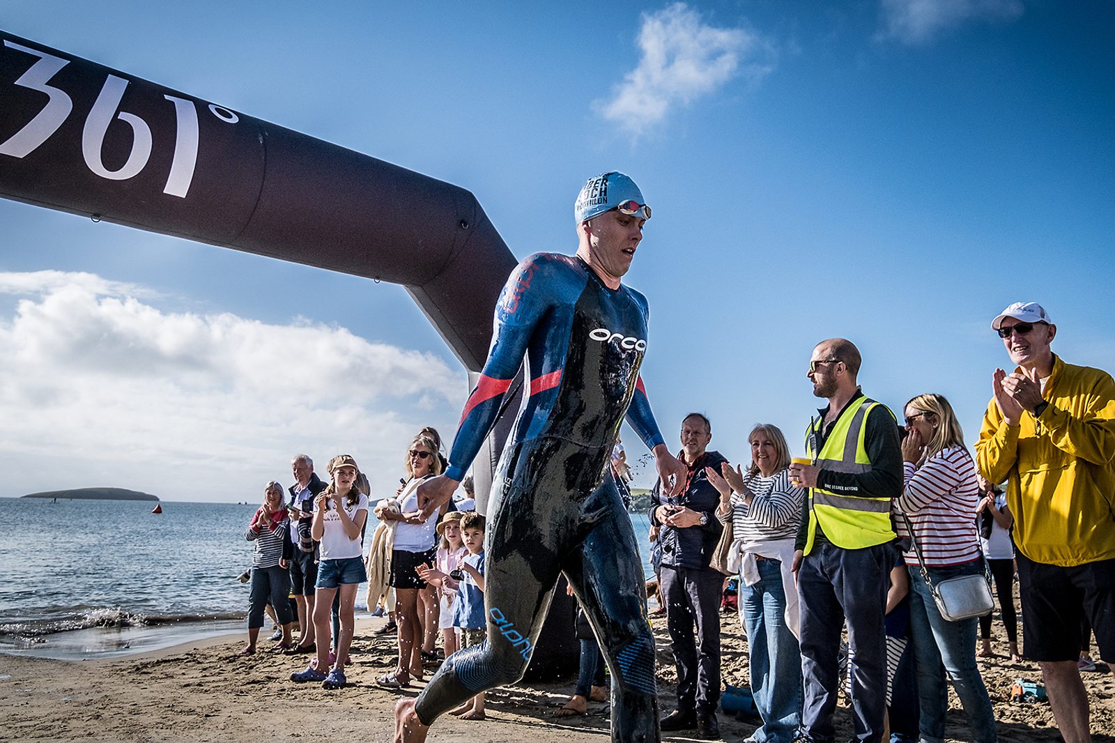 A person in a wetsuit and swim cap emerges from the water and walks on a sandy beach, passing under a black inflatable arch. Spectators line the sides, clapping and cheering under a clear blue sky.