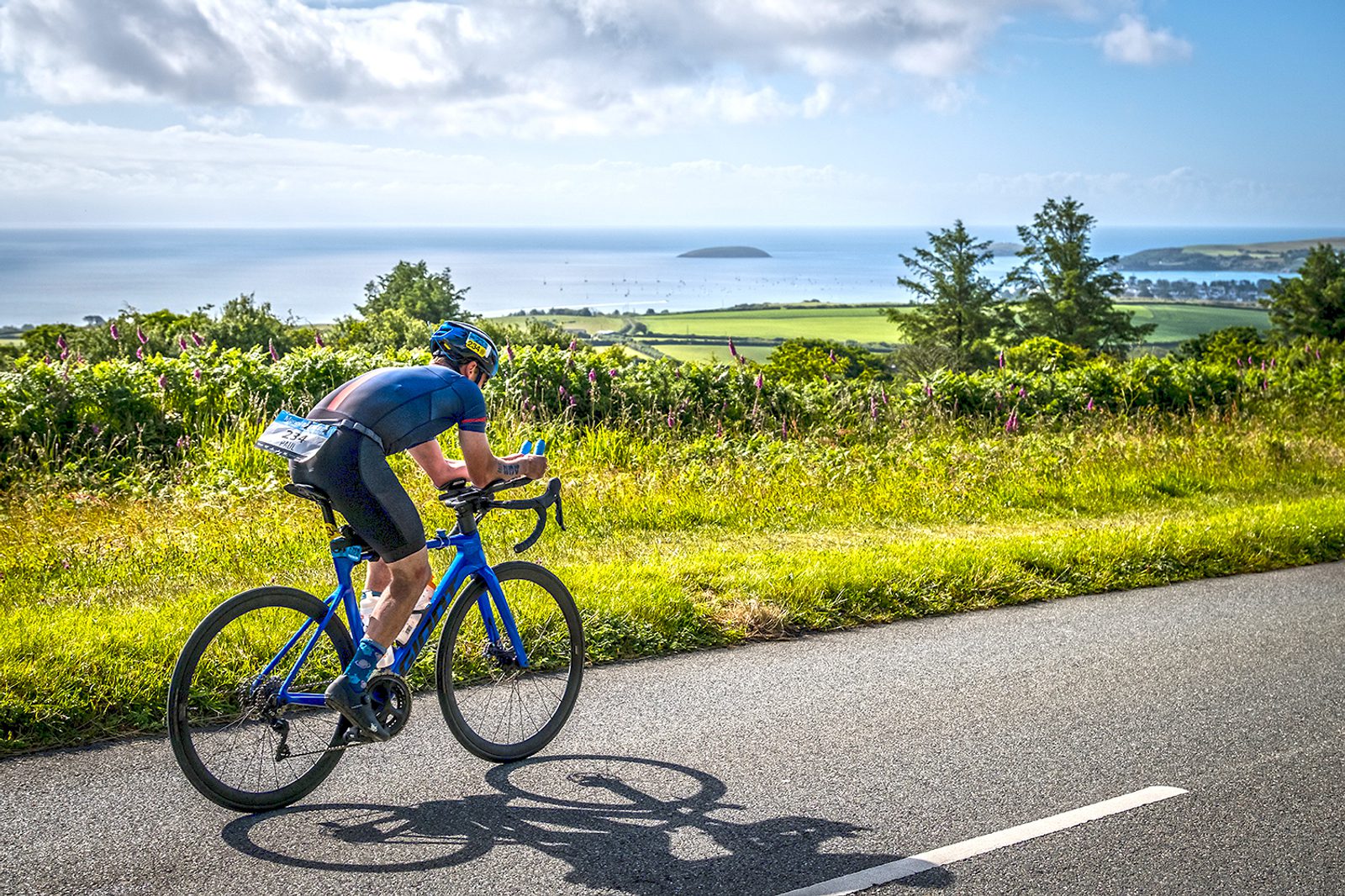 A cyclist in a blue outfit rides a blue road bike on a scenic route with greenery and trees on either side. In the background, there's a panoramic view of the ocean with sailboats and a distant island under a partly cloudy sky.