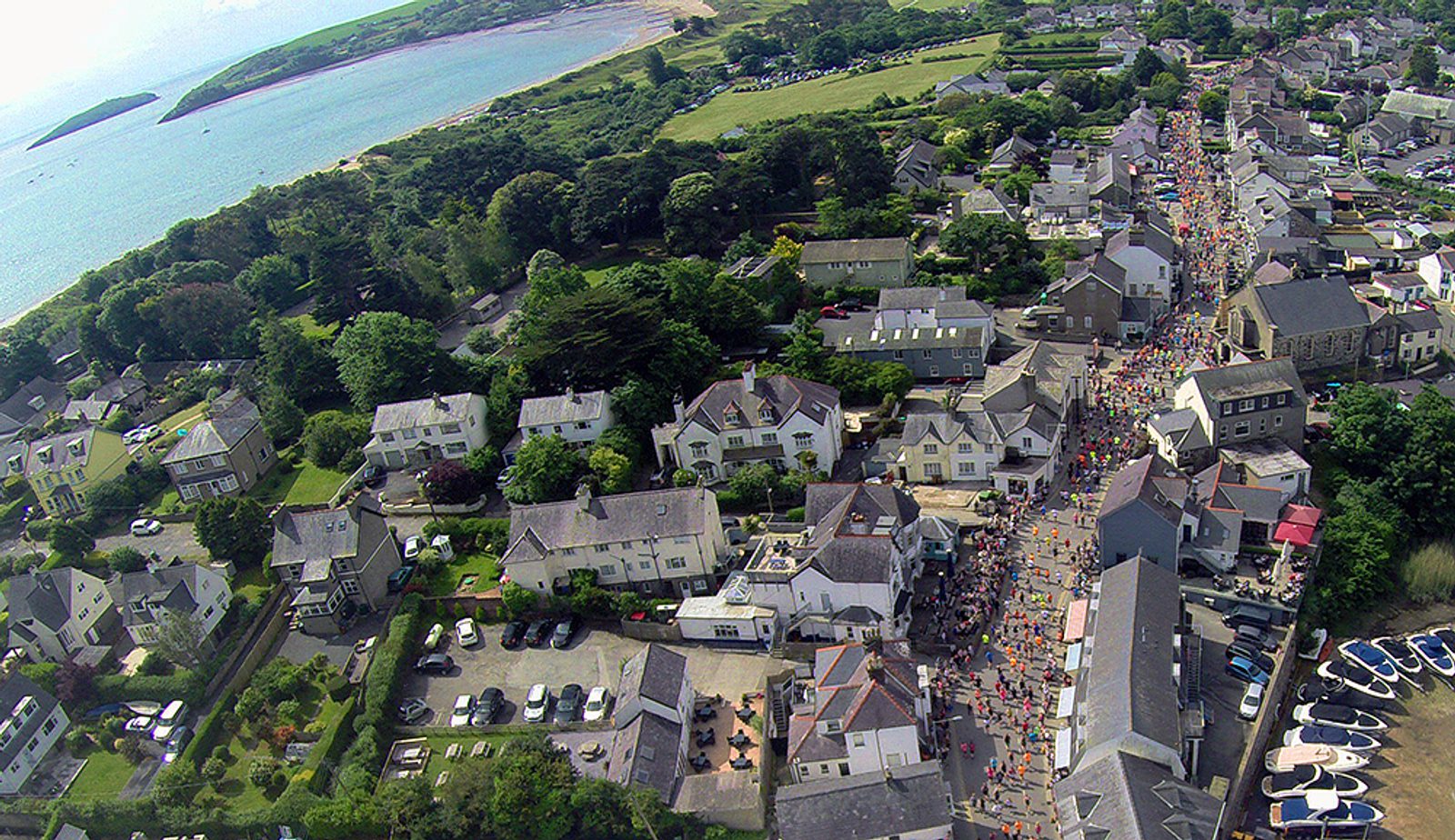 Aerial view of a coastal town with people gathering along a street, possibly for an event or parade. The town is surrounded by greenery and bordered by the sea, with various houses and buildings lining the streets.