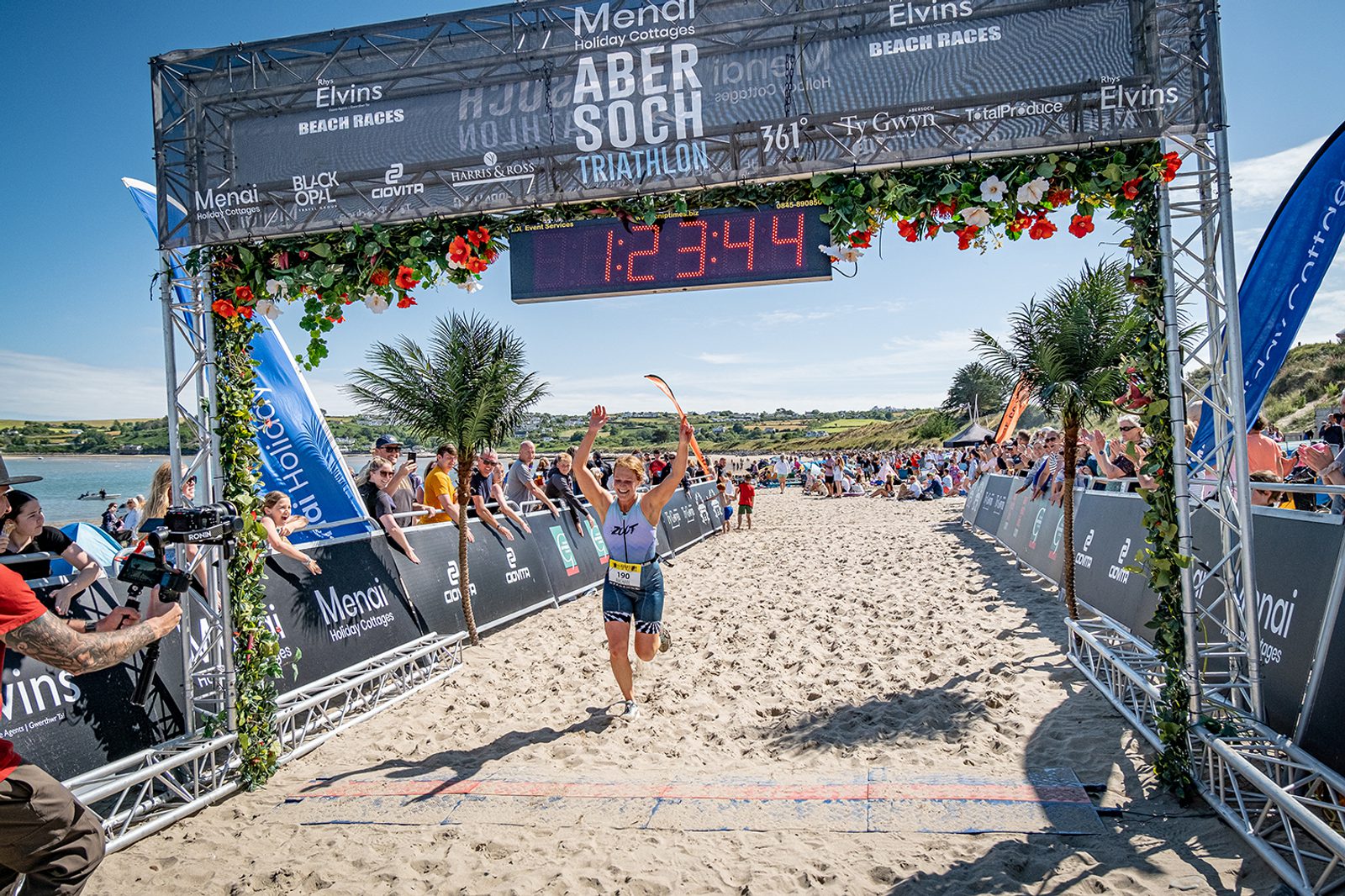 A triathlete crosses the finish line on a sandy beach at the Abersoch Triathlon, with a backdrop of cheering spectators, a timer displaying 1:23:44, and vibrant decorations on the arch. A blue sky and scenic coastline are visible in the background.