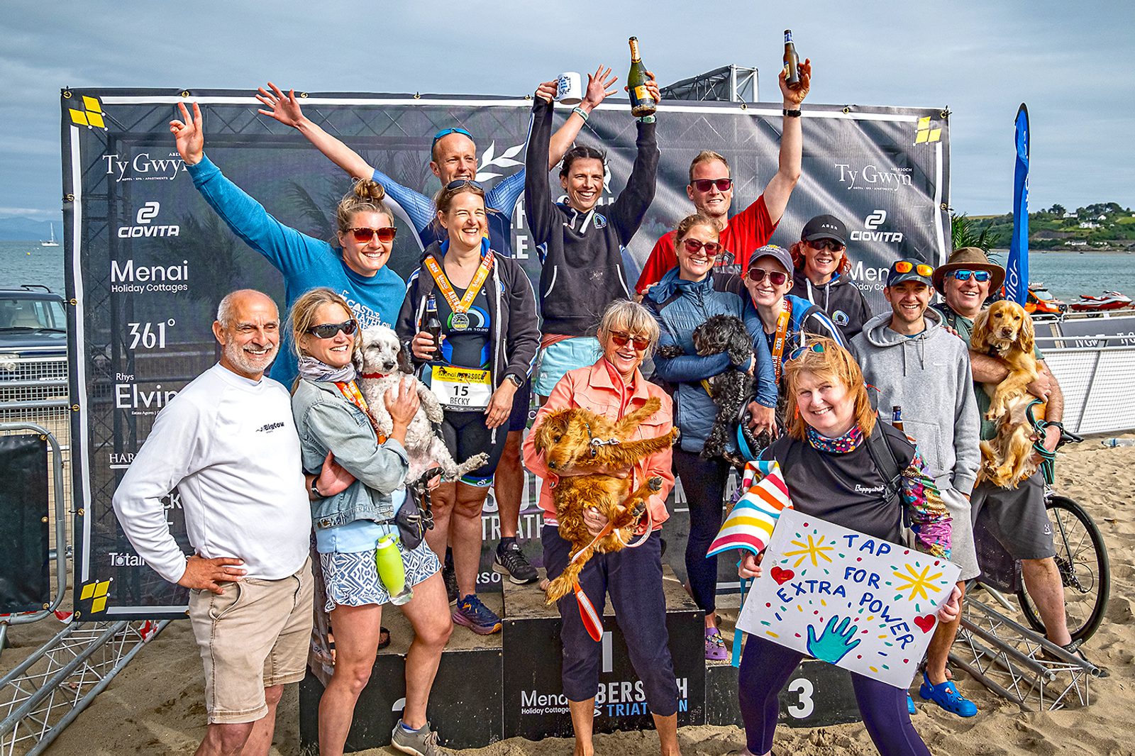 A joyful group of people and several dogs pose together on a sandy beach in front of a race banner. Some hold drinks and one person wears a race medal. Everyone is smiling and celebrating. The atmosphere is festive, with blue skies and decorations in the background.
