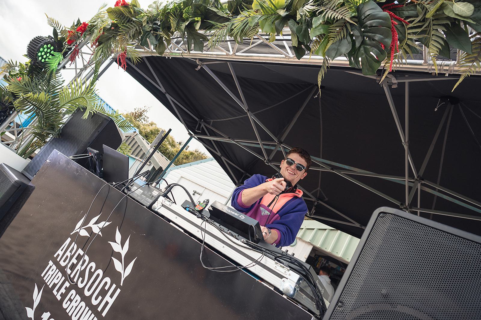 A DJ wearing sunglasses and a colorful jacket is performing on an outdoor stage decorated with tropical plants. The table has a sign reading "Abersoch Triple Crown." Large speakers and equipment are visible.