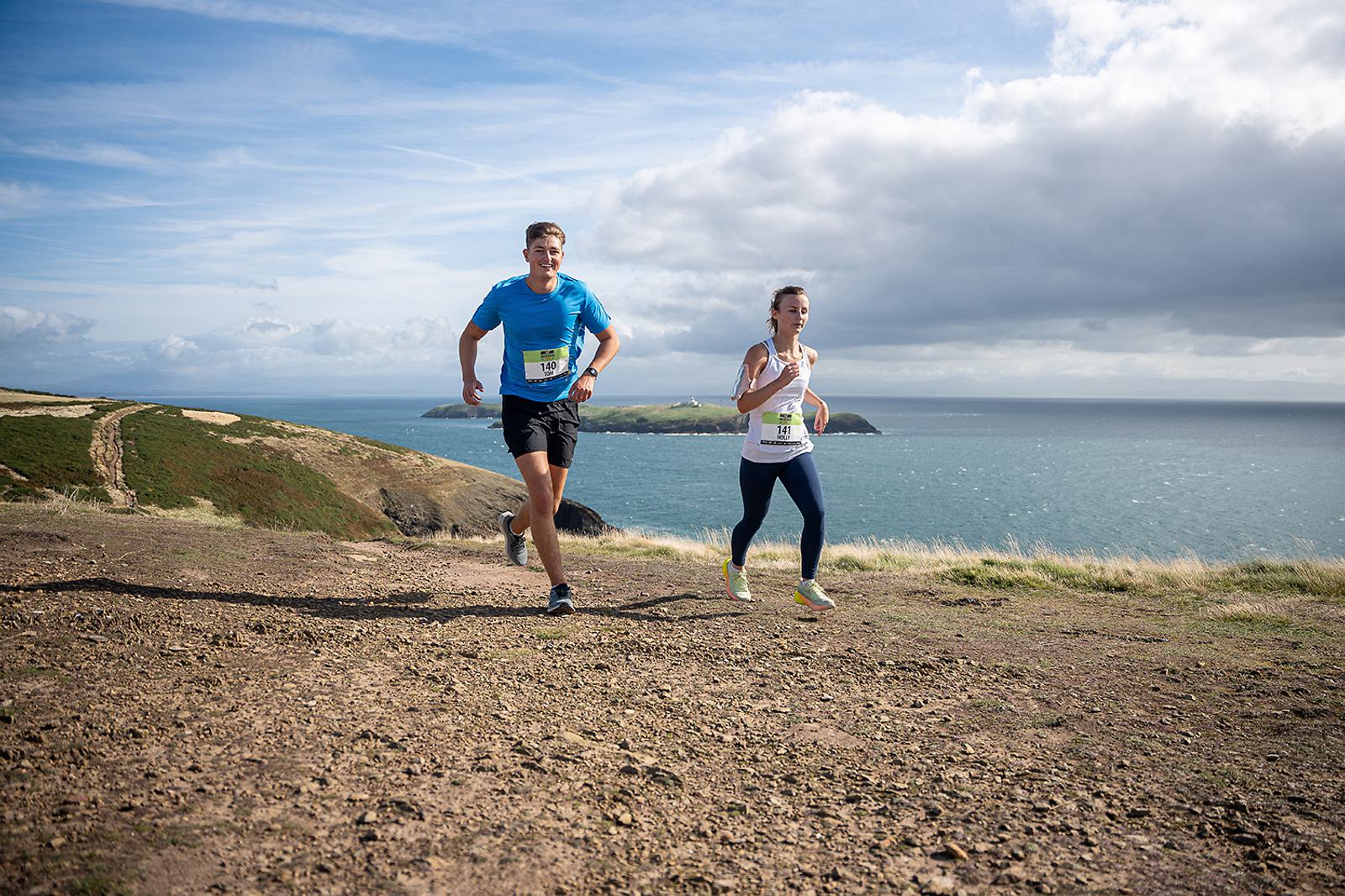 Two people running on a trail near a coastline. The man is wearing a blue shirt and black shorts, and the woman is in a white tank top and leggings. Both have race bibs. The ocean and a partly cloudy sky are in the background.