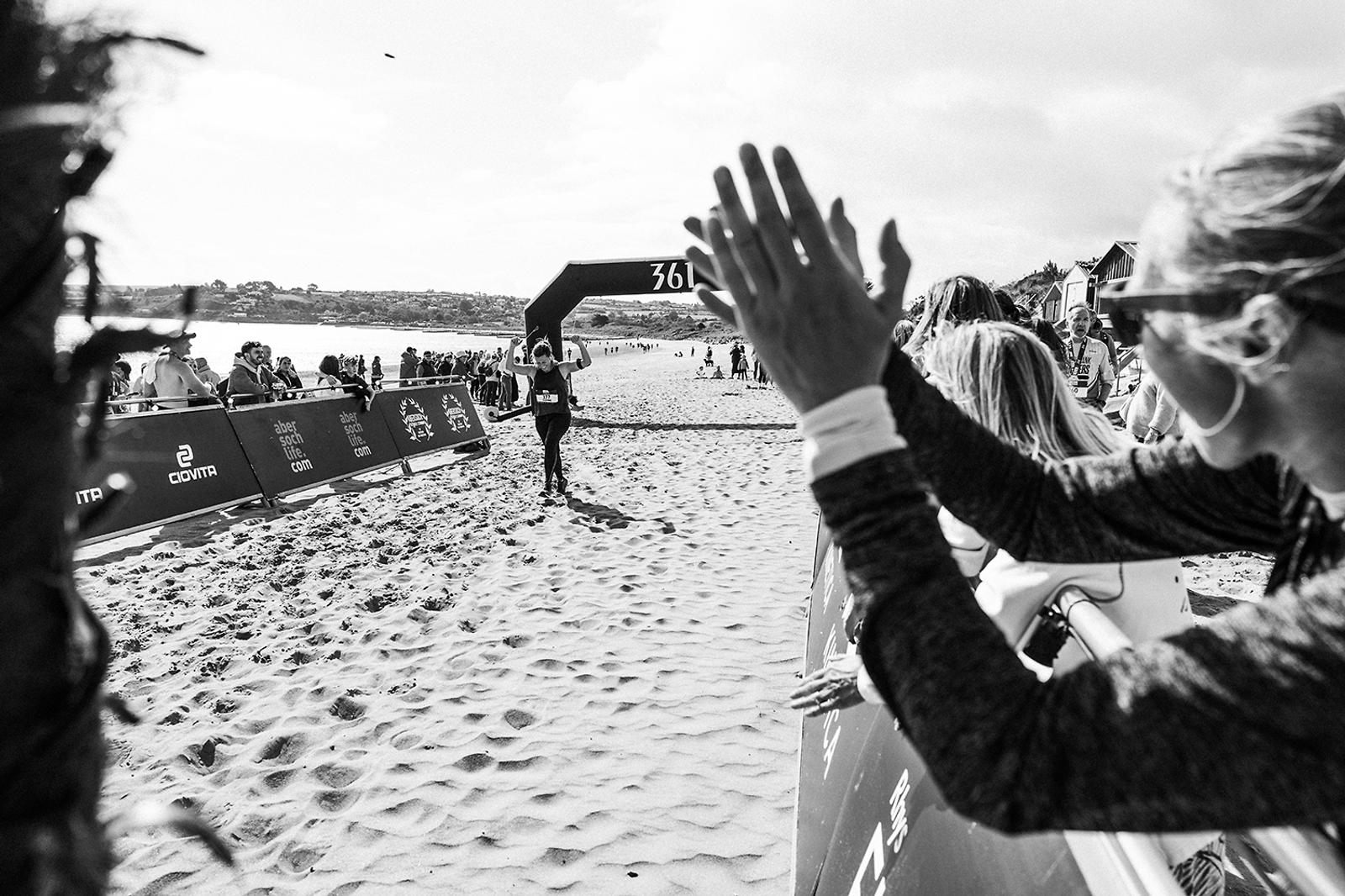 Runner nearing the finish line on a sandy beach race course. Spectators on the right are clapping and cheering. In the background, a large arched banner with race branding is visible. The scene is captured in black and white.