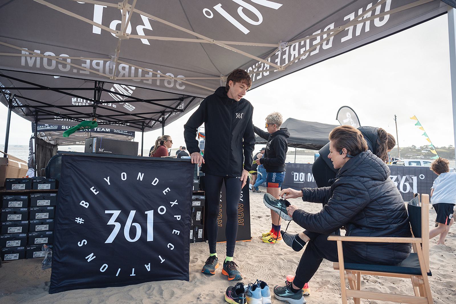 A person stands under a tent wearing athletic gear, while another person sits and helps them try on running shoes. The tent displays "361° Beyond Expectation." Beach and people can be seen in the background.