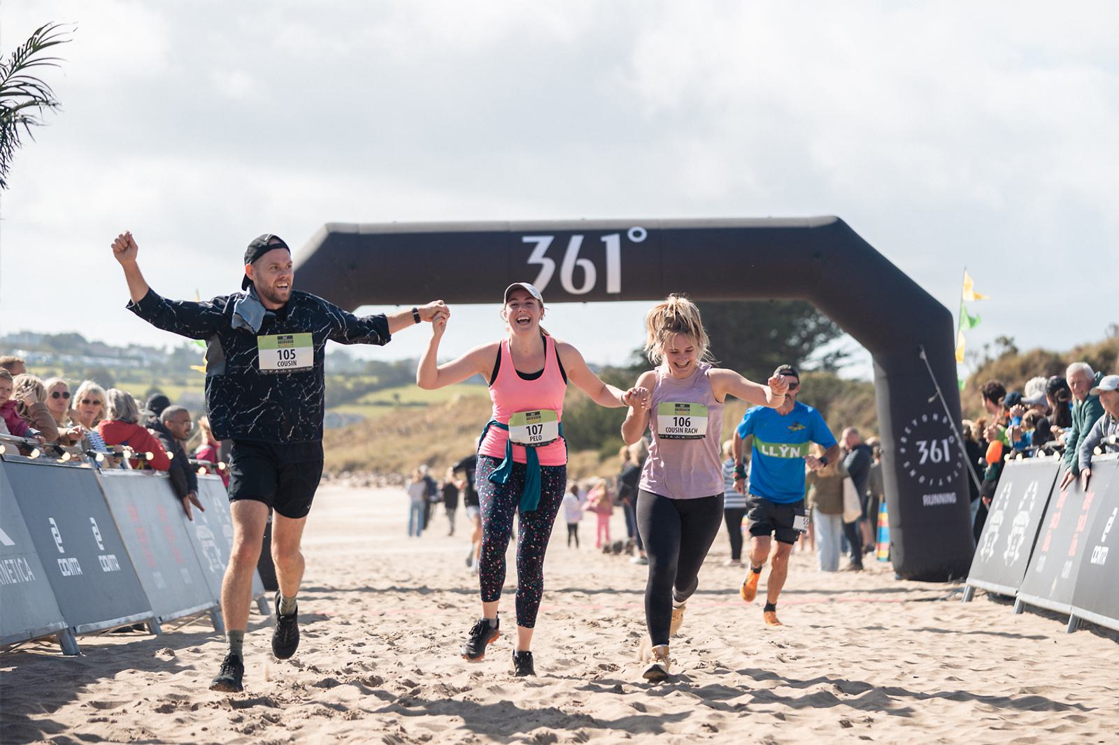 Three runners celebrate as they cross a sandy finish line in a race. The man on the left and two women hold hands, raising them in triumph. Spectators line the sides, and an inflatable arch displays "361°" above them.