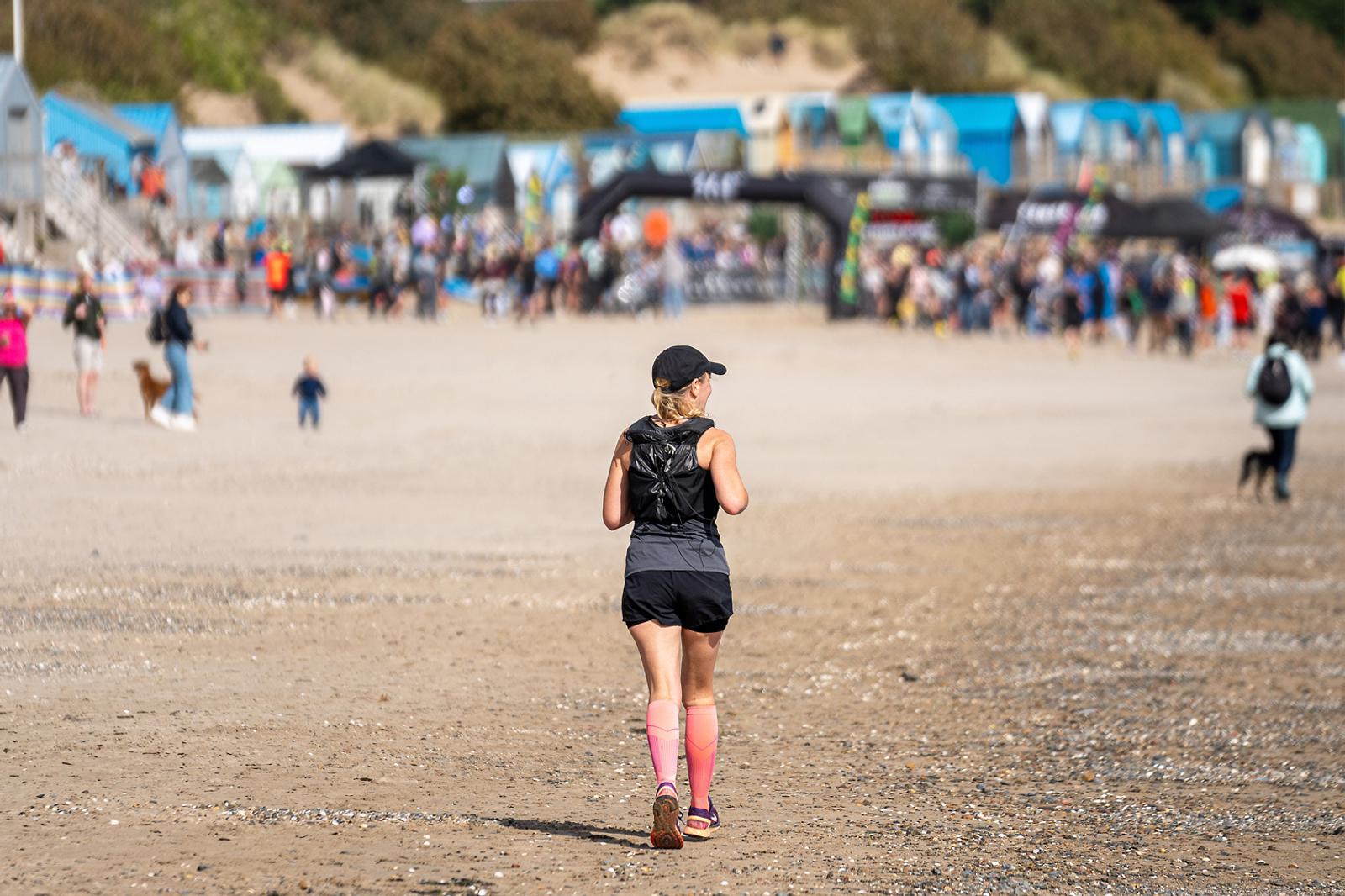 A person wearing a cap, black tank top, shorts, and pink socks is running on a sandy beach. In the background, there are colorful beach huts and a crowd of people near the shoreline under a cloudy sky.