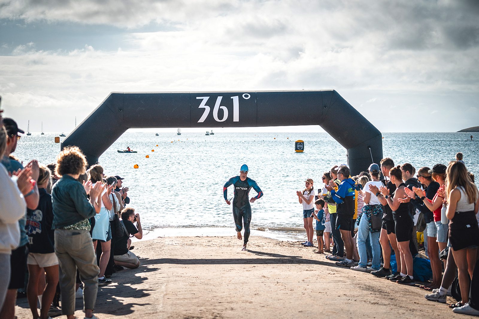 A triathlete wearing a wetsuit and a swim cap runs through an inflatable arch with "361°" written on it. The arch marks the transition area between the swimming and cycling portions of a triathlon. Spectators on both sides cheer as the athlete passes by.