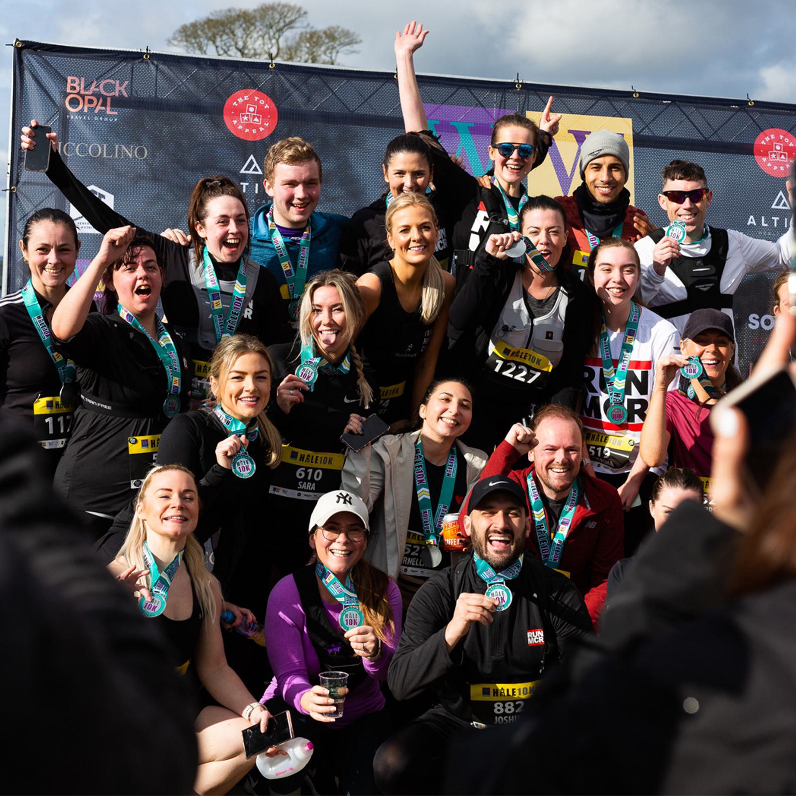 A large, diverse group of smiling runners proudly display their medals and gather for a group photo after completing a race. They're dressed in athletic clothing, some holding drinks, in front of a banner with various logos.