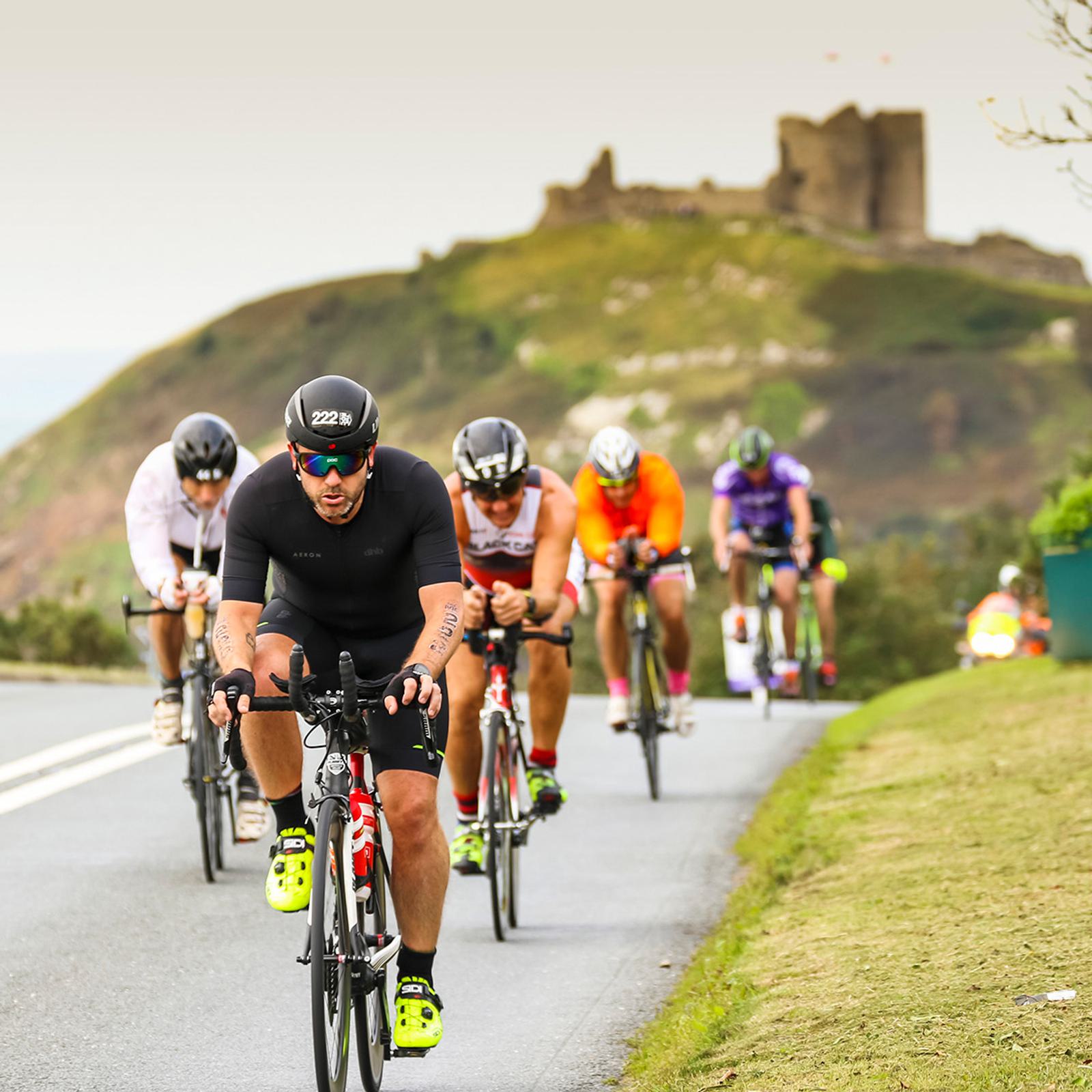 A group of cyclists compete on a hilly road with a blurred castle in the background. The leading cyclist wears black gear and sunglasses, showing intense focus. The other riders follow closely, dressed in various bright colors. The landscape is lush and green.