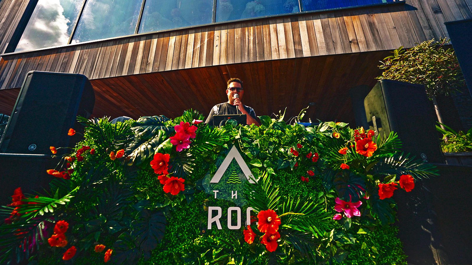 A DJ stands behind a deck decorated with green foliage and vibrant orange and pink flowers. He holds a microphone, wearing sunglasses and a dark shirt. Large black speakers flank him on either side, and a wooden structure with glass windows is in the background.