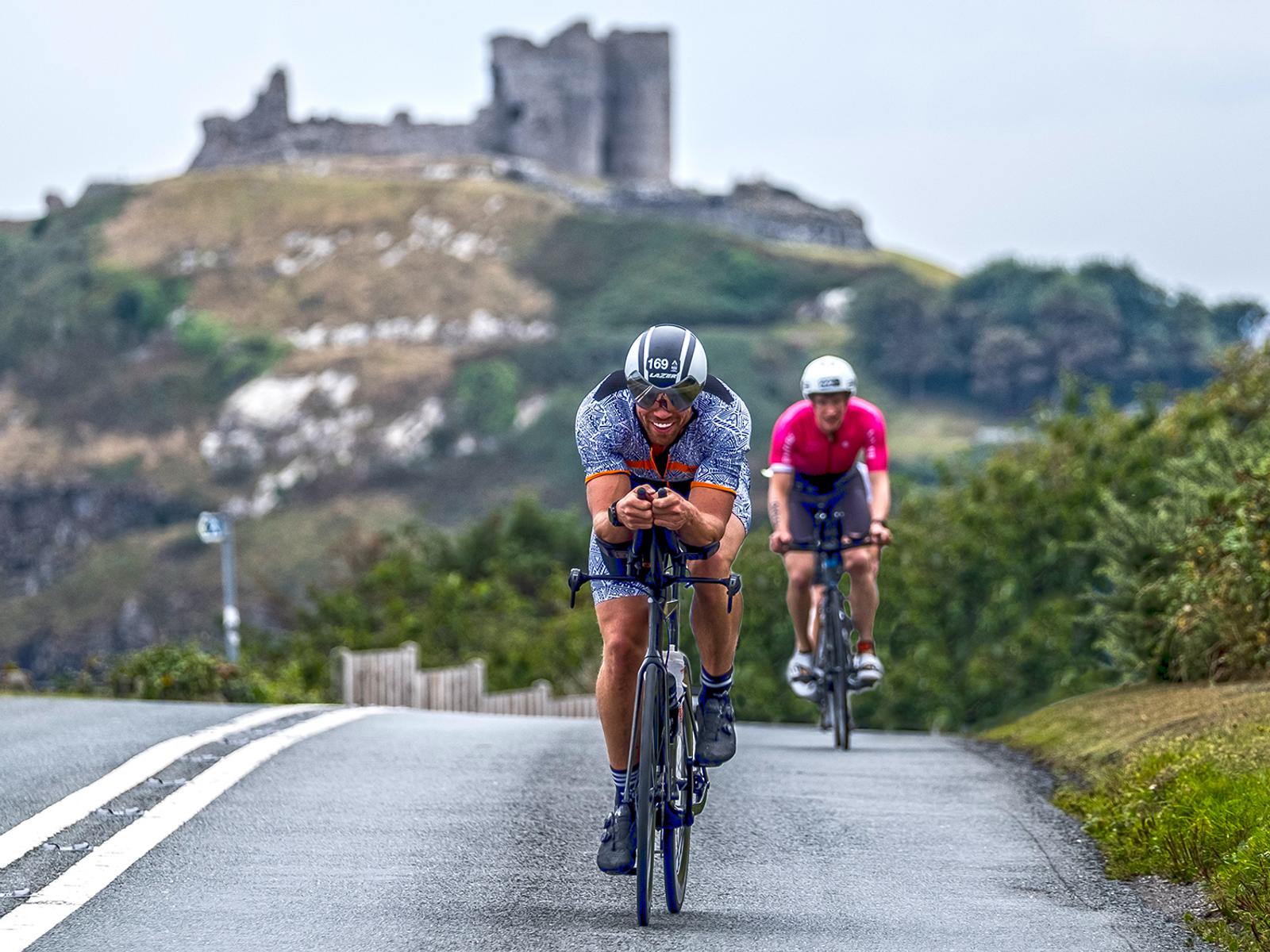 Two cyclists are riding on a road with white lines. The leading cyclist is wearing a blue kit and a helmet, hunched over the handlebars. The second cyclist in a pink kit follows behind. A historic castle on a hilltop is in the background under a cloudy sky.