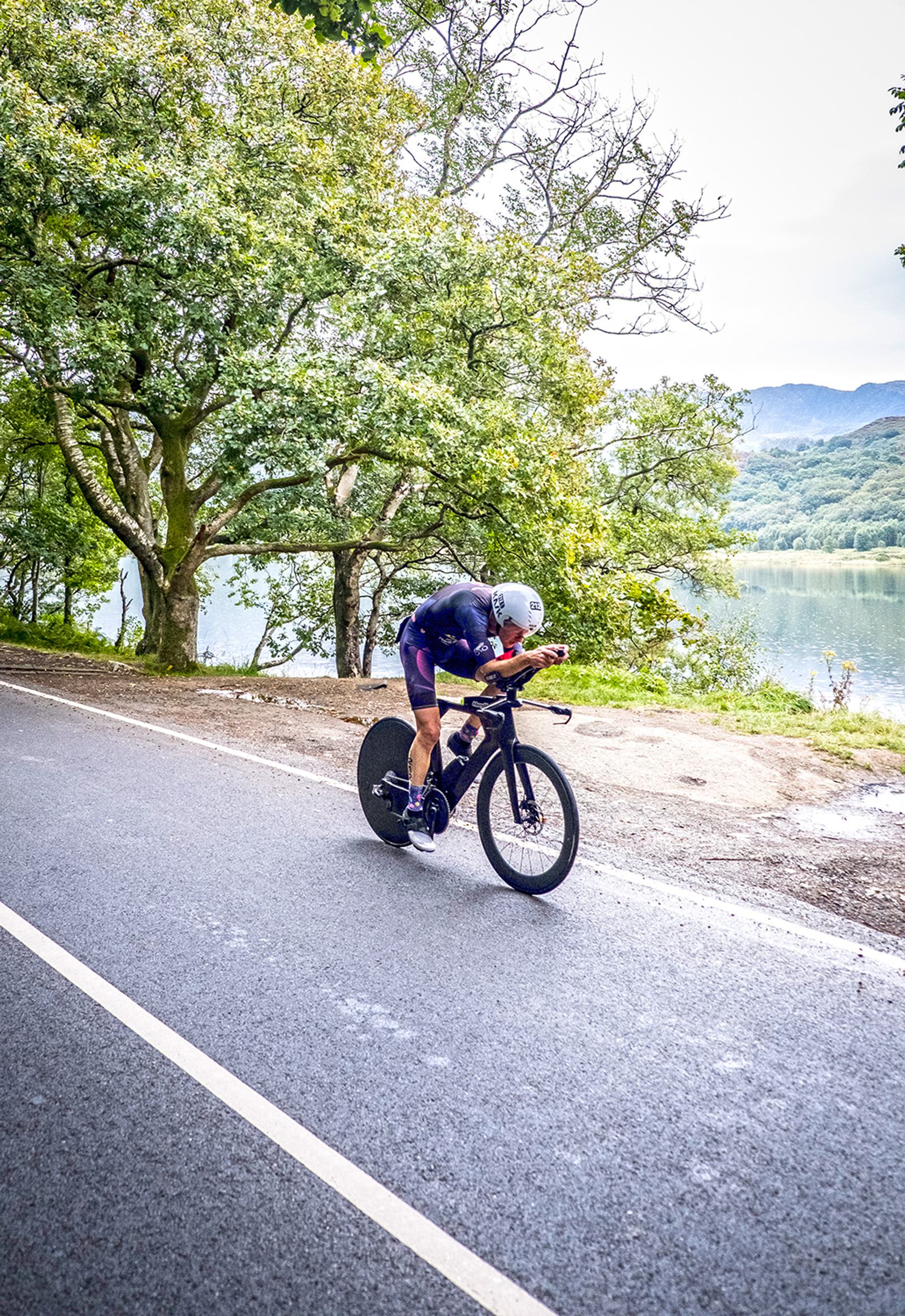 A cyclist in aerodynamic gear rides a bicycle down a paved road lined with trees and a lake in the background. The cyclist is bent forward, displaying intense focus, with lush greenery and distant hills visible under a cloudy sky.