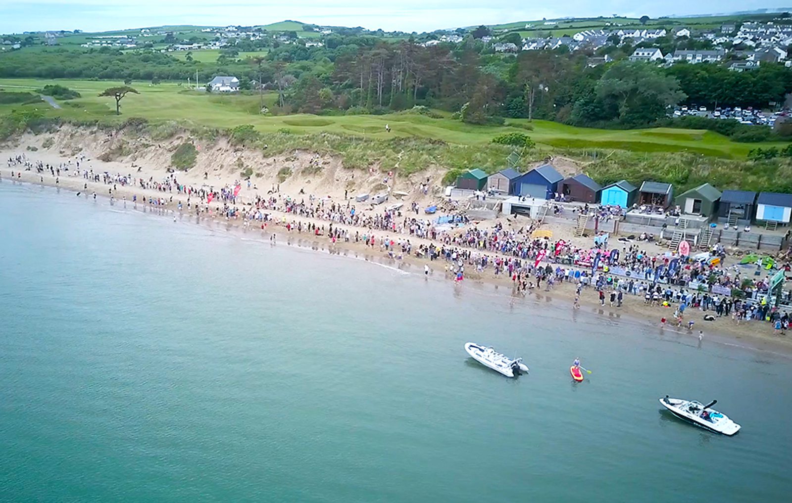 Aerial view of a crowded beach with people along the shore, several boats in the water, and a row of colorful beach huts in the background. The scene is surrounded by grassy dunes and lush greenery with houses visible in the distance.