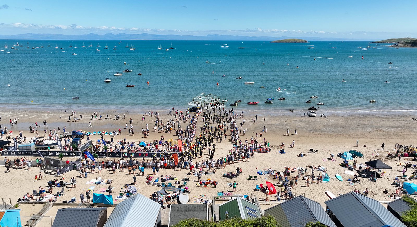 A crowded beach on a sunny day with people gathering around a pier. Several tents and beach huts are visible, along with boats and paddleboarders on the water. The background features sailboats and a distant island under a clear blue sky.