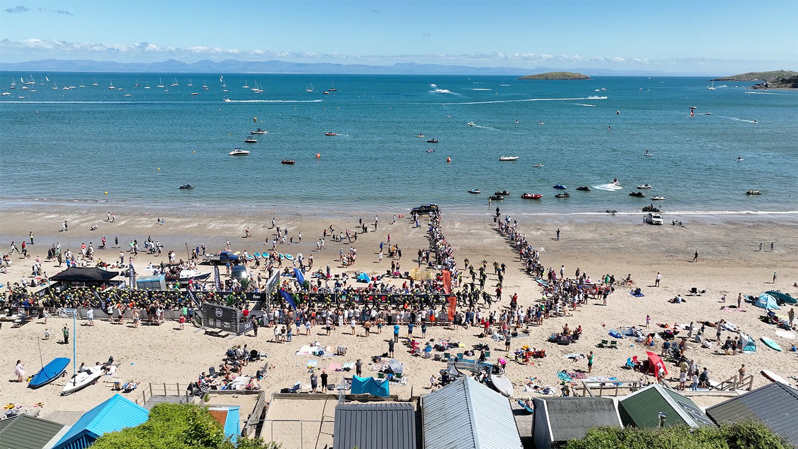 A crowded beach scene with many people gathered near the shoreline. Numerous pop-up tents and structures are set up on the sand. Boats and sailboats are visible in the water, and hills are seen on the horizon under a clear blue sky.