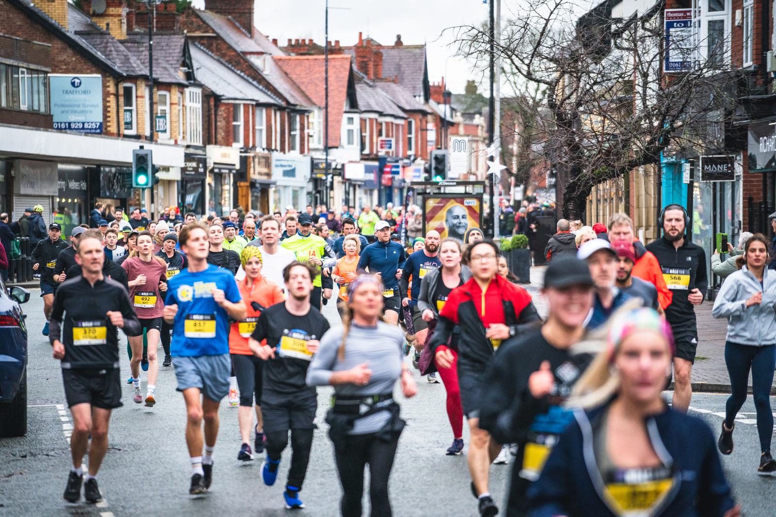 A group of runners participate in a race on a street lined with shops and spectators. The runners, dressed in various athletic gear, race forward with determined expressions. A green traffic light is visible, and the weather appears overcast.