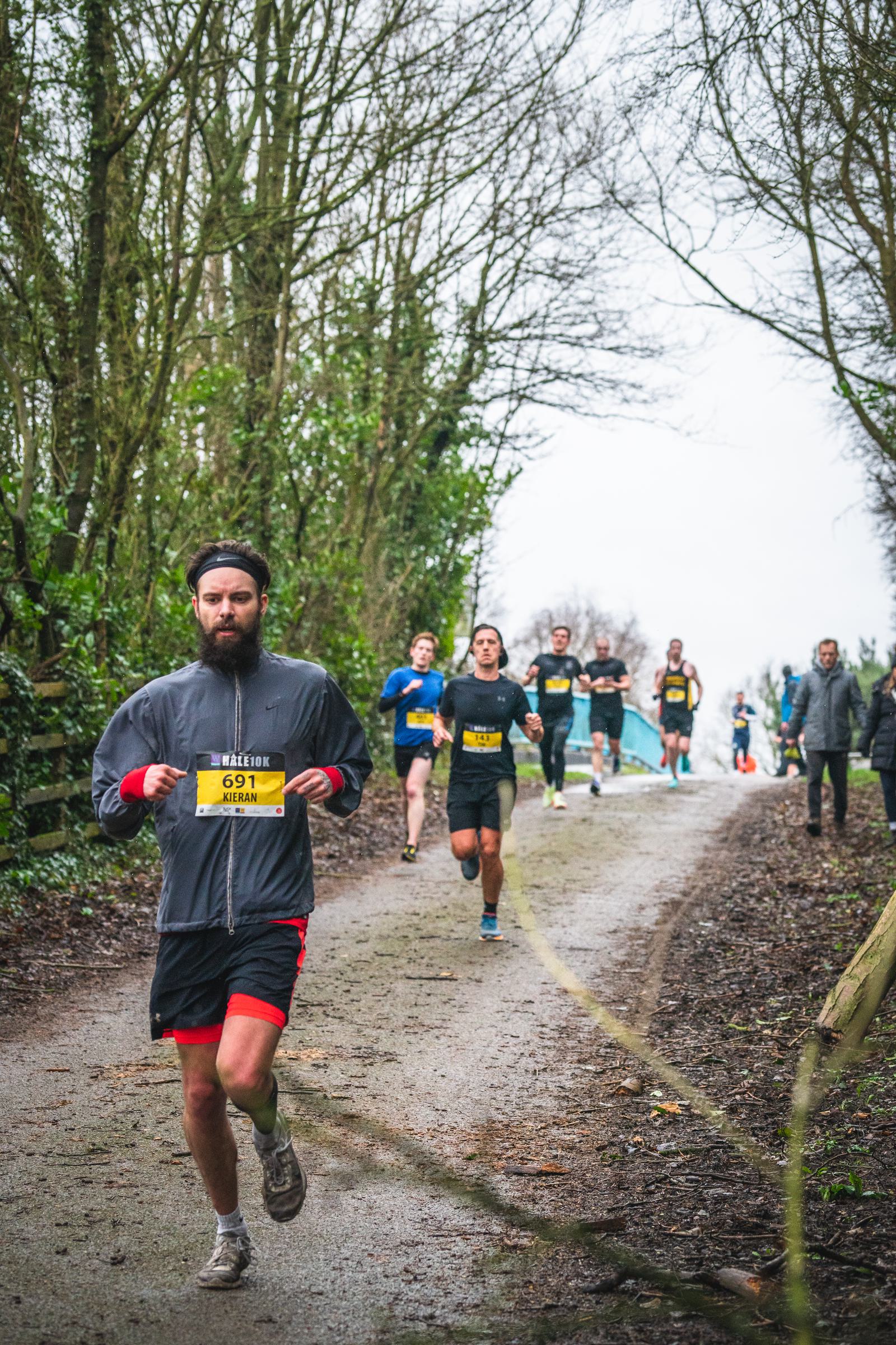 A group of people running on a forest trail during a race. The runner in the foreground wears a grey jacket, black leggings, and shorts, with a yellow race number 691. Trees line both sides of the path, and more runners are visible behind, with a cloudy sky overhead.