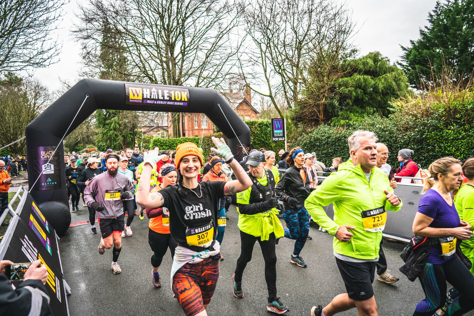 A group of runners is participating in a race, with some smiling and waving at the camera. They are running under a "Wilmslow 10K" inflatable arch on a tree-lined street. The runners are dressed in colorful athletic gear and numbered bibs. The atmosphere is energetic and lively.