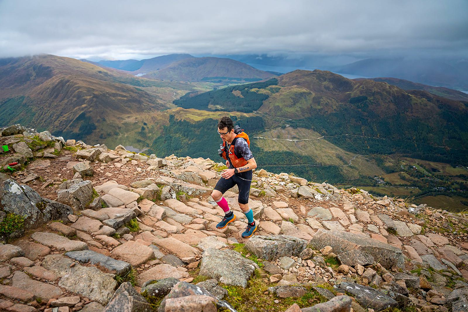 A person running uphill on a rocky mountain trail, wearing a tank top and shorts. They have colorful compression socks and a backpack. The background features expansive green hills and a cloudy sky.