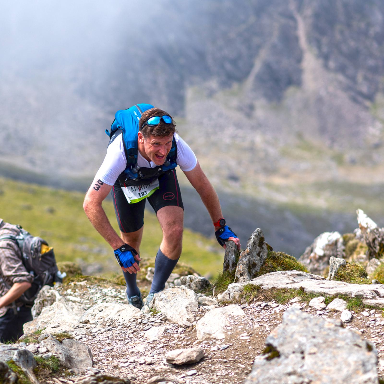 A man wearing a white shirt, blue backpack, and knee-length socks is climbing up a rocky mountain trail. He appears to be struggling but determined, with a misty, mountainous background. Another climber is partially visible in the background.