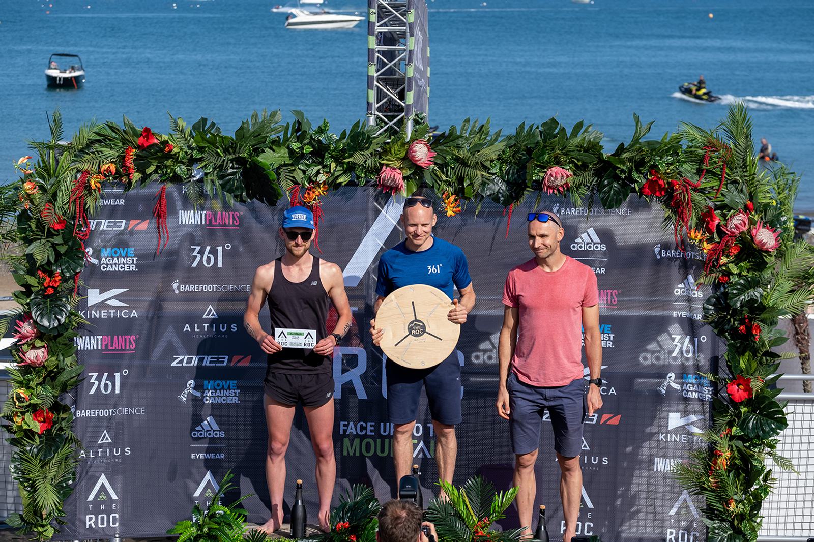 Three men stand on a podium at a waterfront event, surrounded by greenery and sponsor logos. The man in the center holds a trophy shaped like a wooden shield, while the man on the left holds a plaque. In the background, boats are visible on the water.