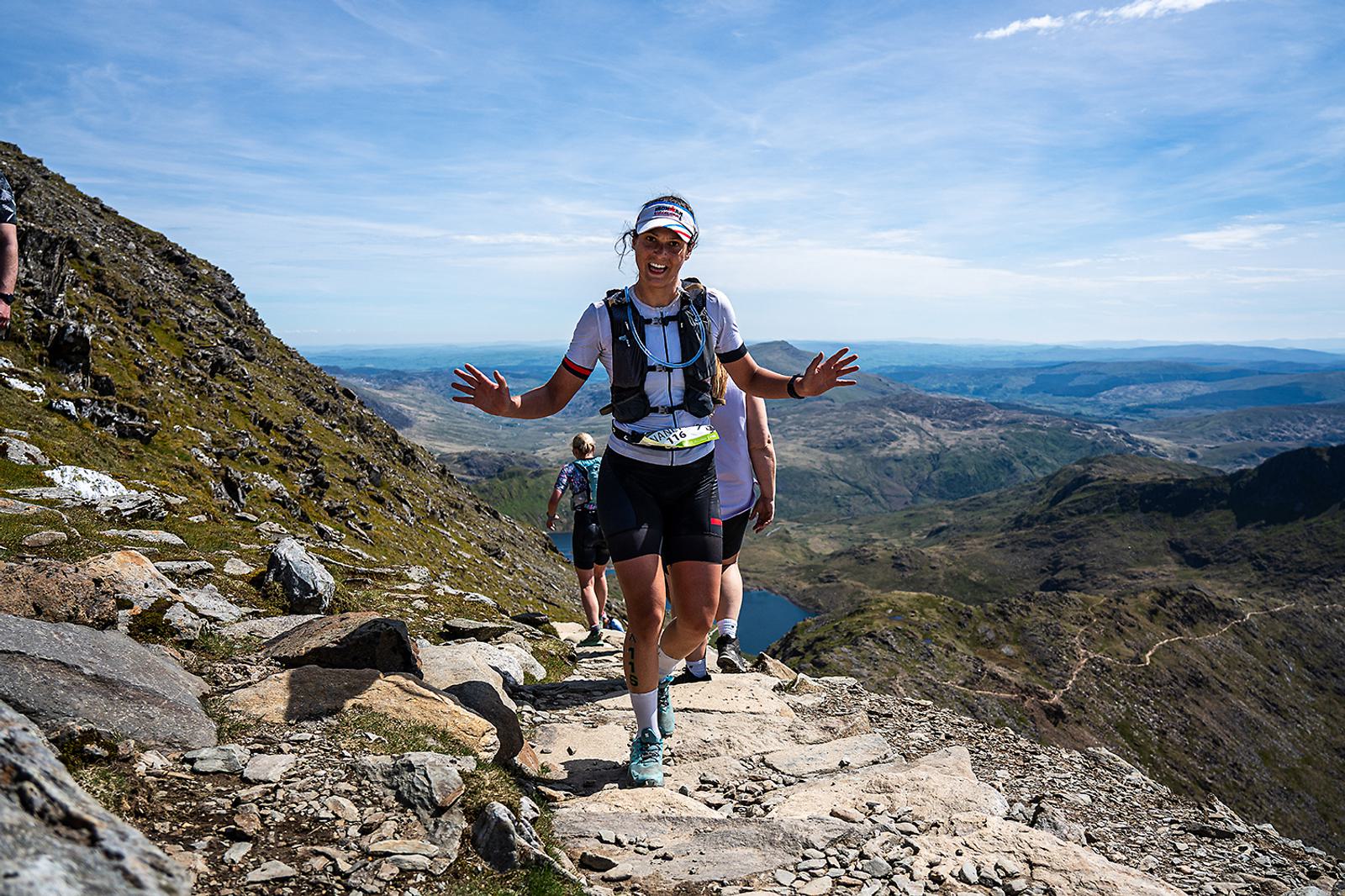 A runner wearing a hydration pack and race bib smiles and gestures with hands raised while ascending a rocky mountain trail. The background features expansive views of a mountainous landscape and a bright blue sky.