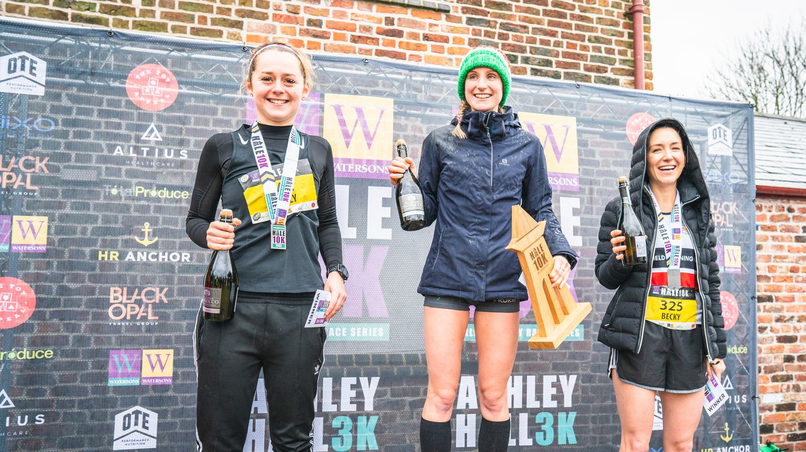 Three female runners stand on a podium after a race, smiling and holding bottles of champagne. The central athlete, holding a large wooden trophy, is on the highest step of the podium. They are in front of a backdrop with sponsors' logos.