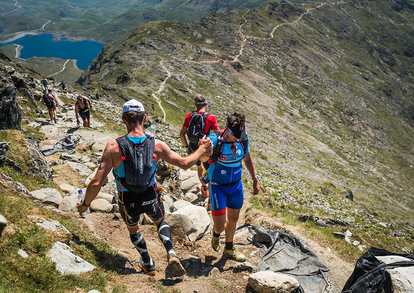 Runners navigate a rocky mountain trail with a scenic lake in the background. The terrain is rugged, and some runners are wearing athletic gear and hydration packs. The path is steep and surrounded by lush green hills and rocky outcrops under a clear sky.