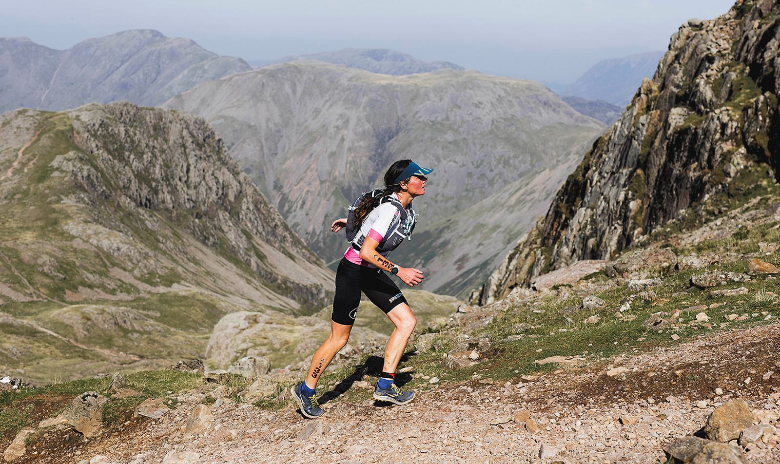 A person in athletic gear and sunglasses runs up a rocky mountain trail, surrounded by expansive mountain views under a clear sky. The scenery includes rugged peaks and a distant, hazy horizon.