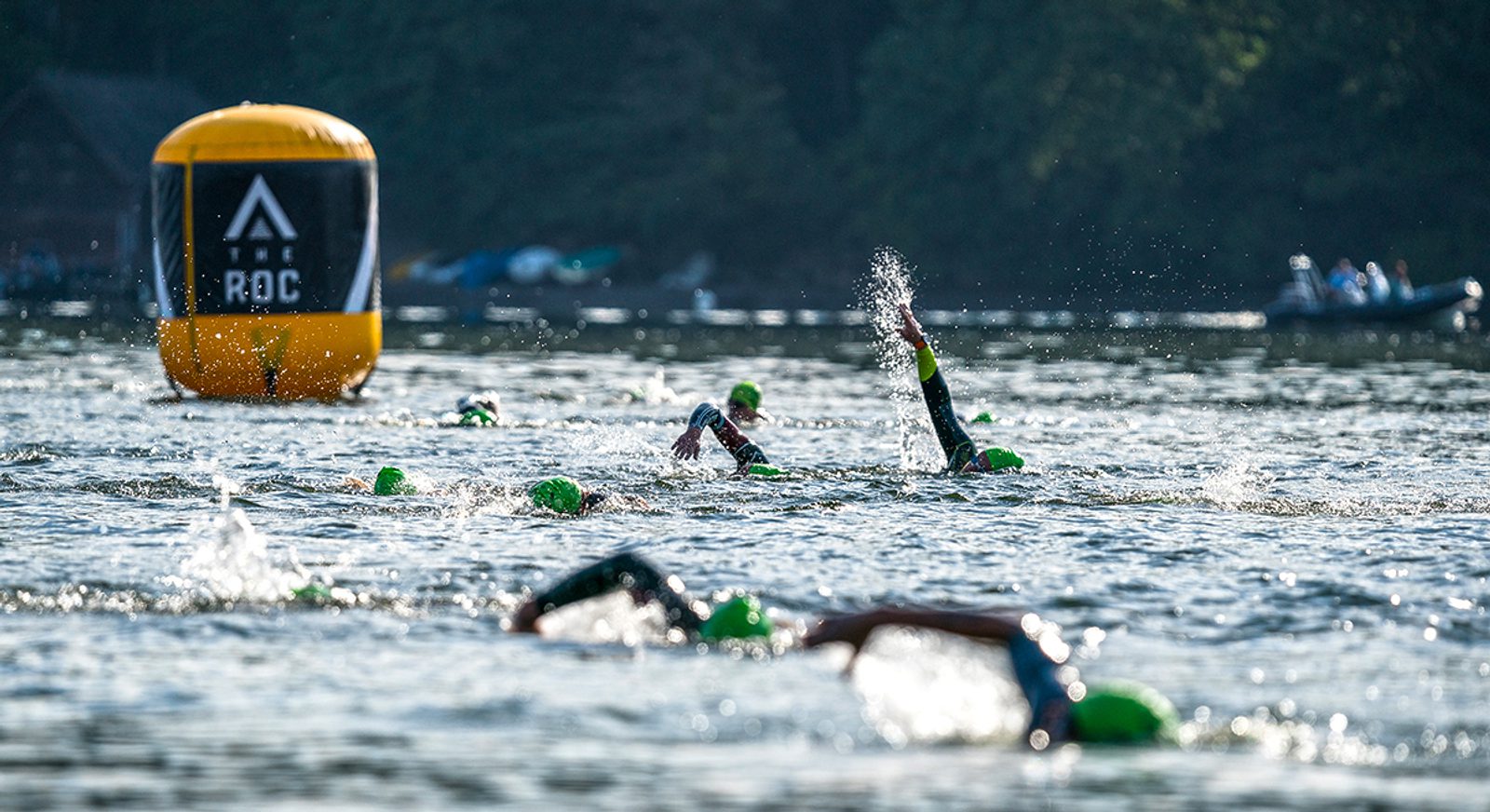 Swimmers wearing green caps compete in an open water race. They are moving towards a large yellow buoy in a lake. The background shows trees, people on boats, and a dock area. The water is slightly choppy, with sunlight glinting off the surface.