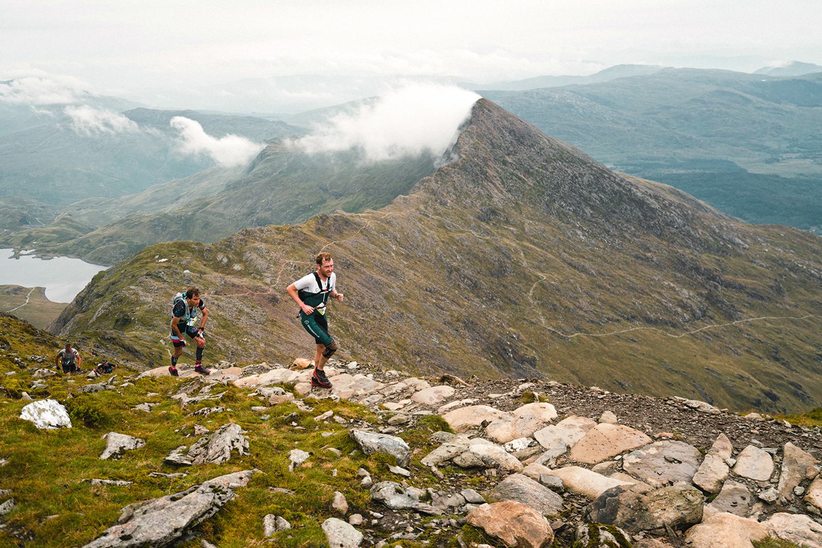 Three hikers are ascending a rocky mountain trail with expansive, misty landscapes in the background. One hiker is in the lead, while two others follow closely behind. A sharp, prominent peak and patches of greenery are visible in the distance.