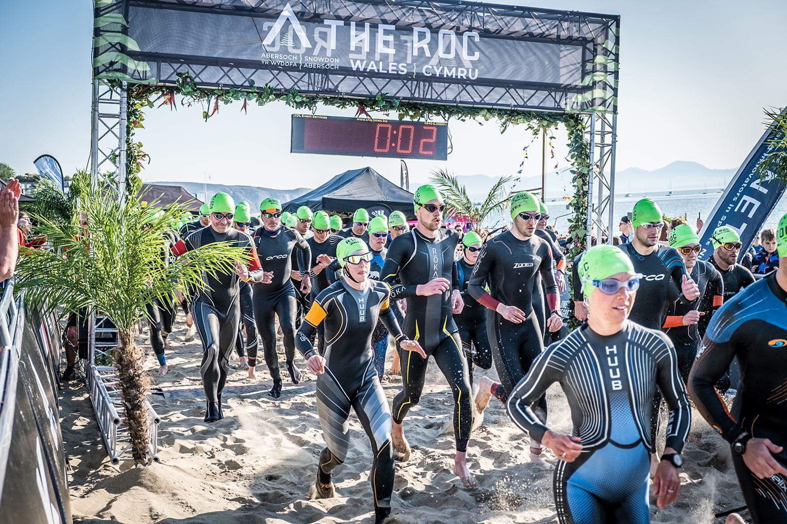 Athletes in wetsuits and green swim caps begin a triathlon on a sandy beach, running towards the water under a starting banner. Palm fronds line the path, and a digital clock displays the time as 00:02.