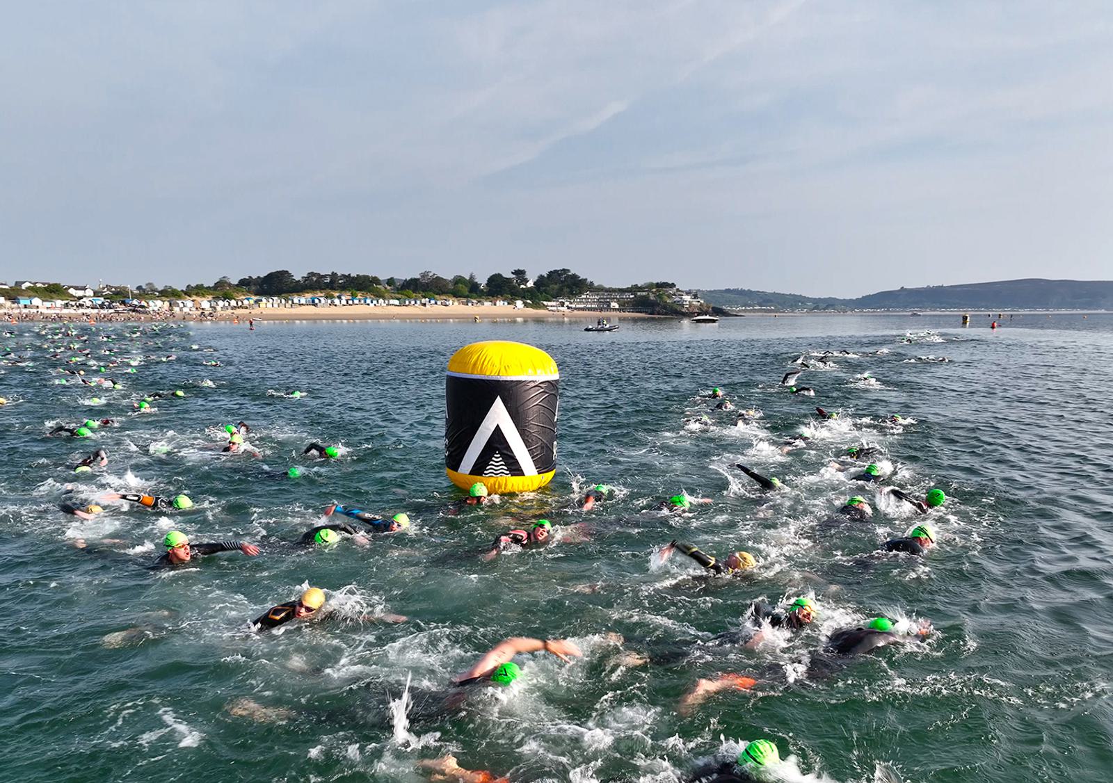 A large group of swimmers wearing green swim caps participate in an open water race. They swim around a yellow marker buoy in a vast body of water, with a distant shoreline and cloudy sky in the background.