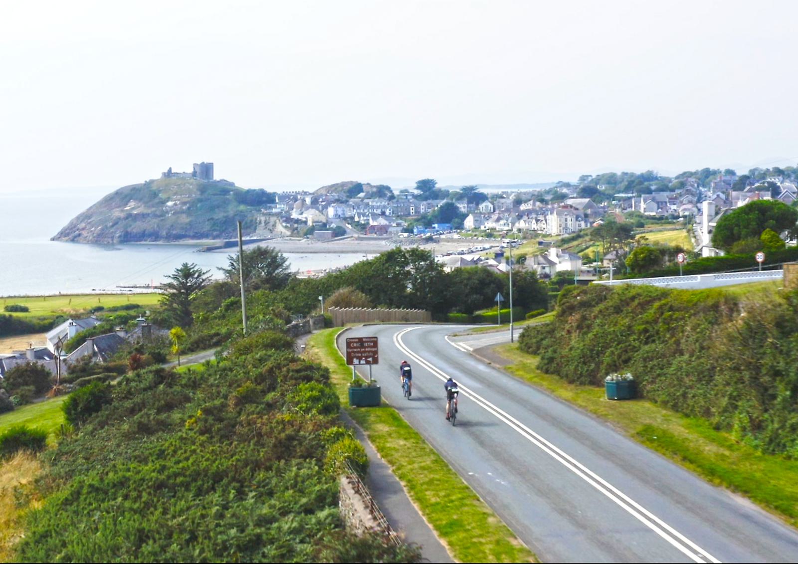 Three cyclists ride along a winding road through a lush coastal landscape. In the distance, a seaside town is nestled near the shore, with a hilltop castle overlooking the scene. The sky is partly cloudy, creating a serene atmosphere.