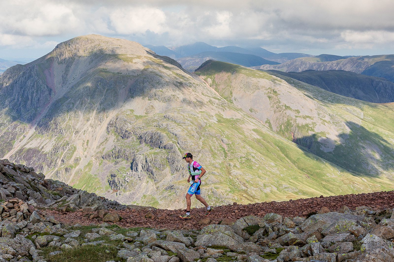 A person wearing athletic gear, including a hat and a backpack, is running on a rocky terrain with a mountainous landscape in the background. The distant mountains are partially shrouded by clouds on a sunny day.
