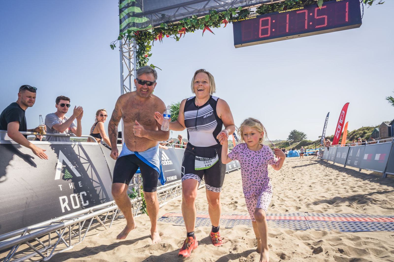 A woman wearing a triathlon suit runs towards the finish line on a sandy beach, holding hands with a man in blue swim trunks and a young girl in a pink dress with polka dots. A digital timer above shows 8:17:51. Spectators cheer and clap in the background.