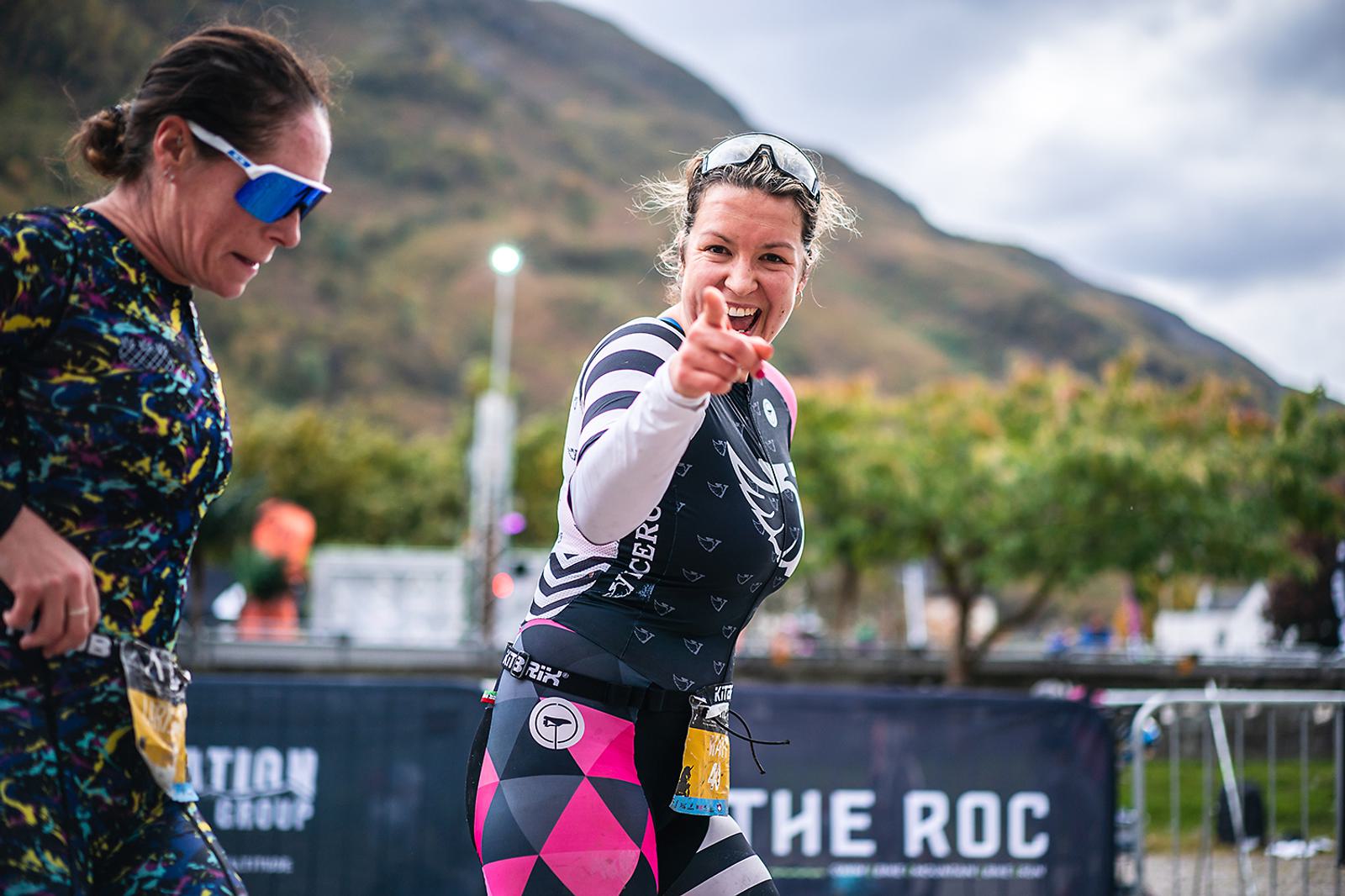Two women in athletic gear participate in a race outdoors. One woman smiles and points towards the camera. Mountains and greenery form the backdrop, with a banner visible in the background.