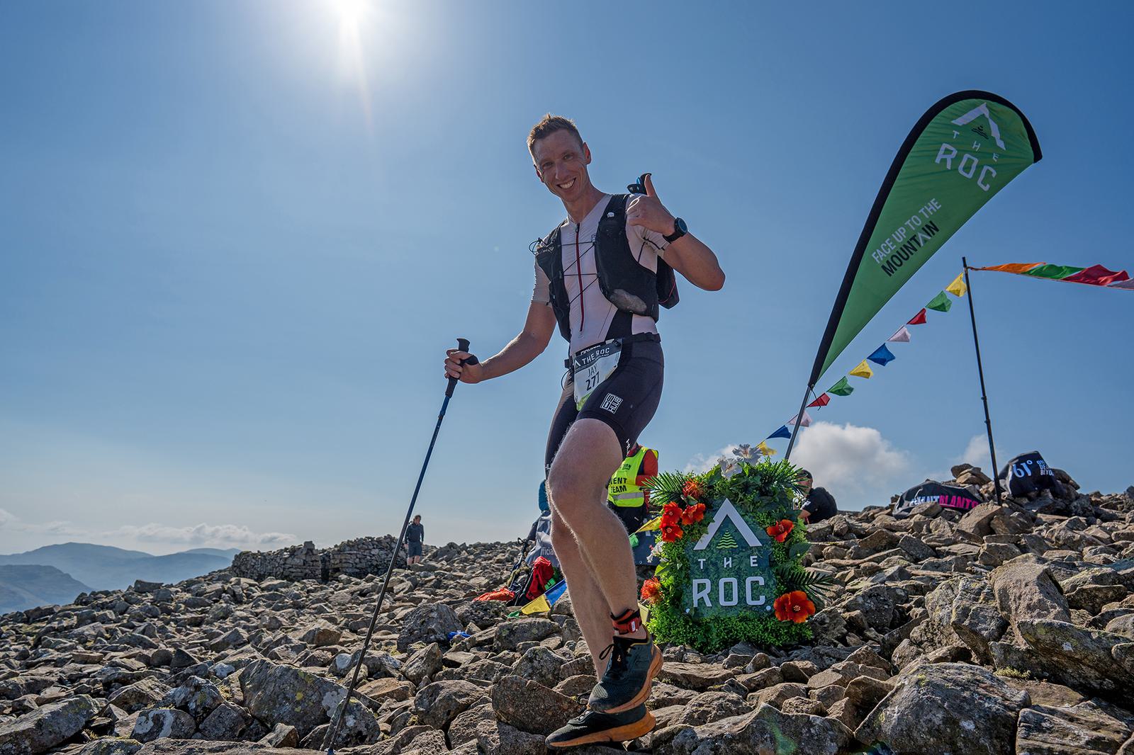 A person wearing athletic gear and holding a hiking pole stands on a rocky mountain peak under a clear blue sky. They are smiling and gesturing thumbs up, with a flag that reads "The Roc" in the background.