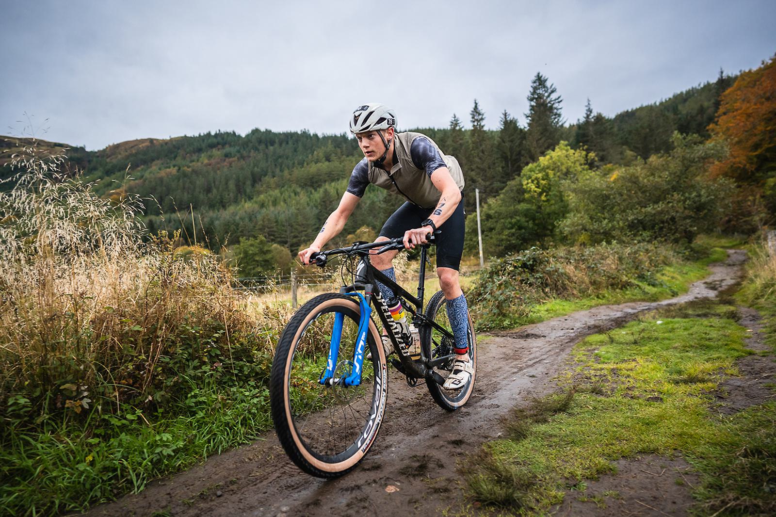 A person wearing a helmet and cycling gear is riding a mountain bike on a muddy trail through a forested area. The background is lush with greenery and trees under an overcast sky.