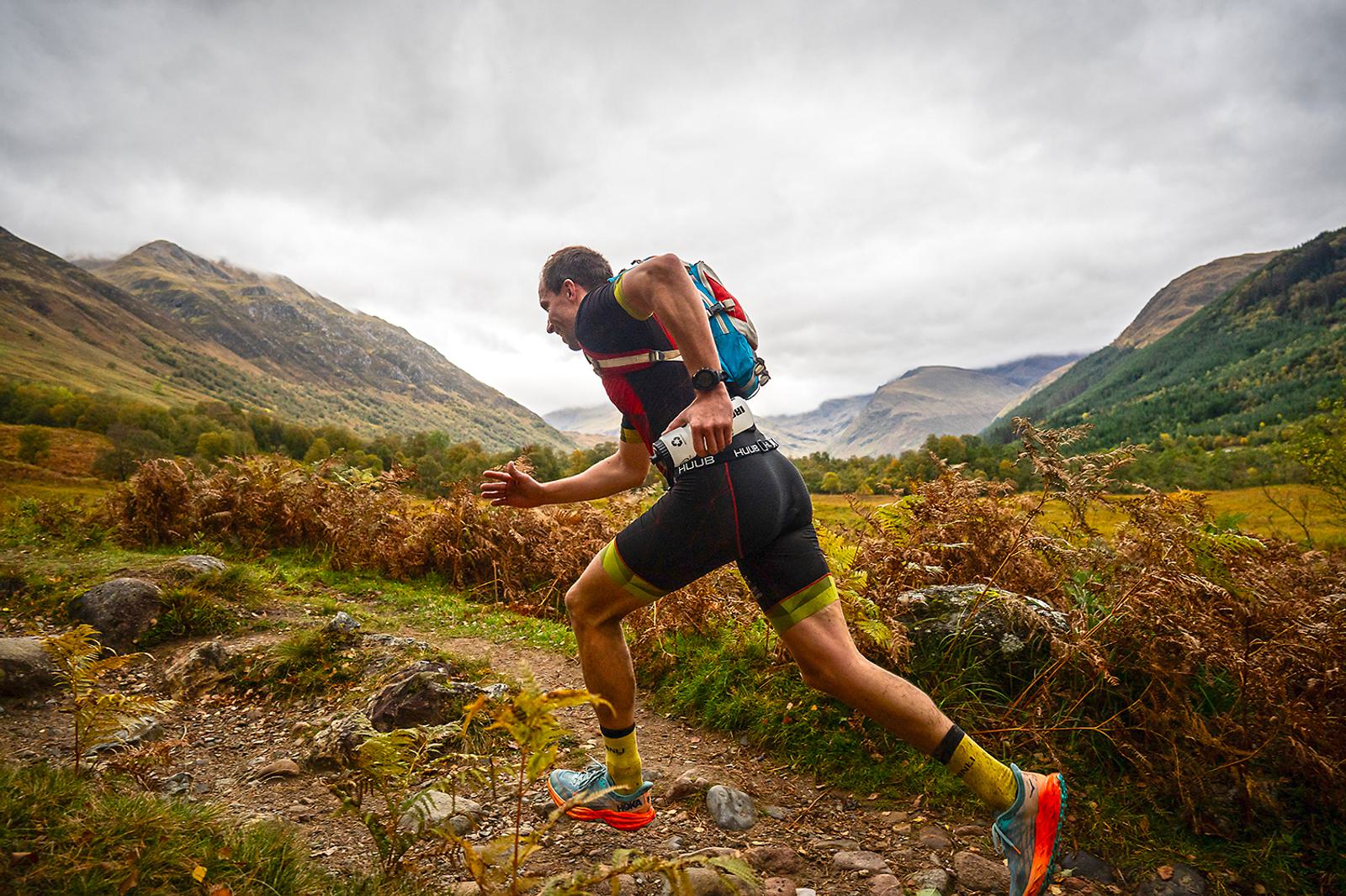 A man in athletic gear runs on a rocky trail through a scenic mountainous landscape. He wears a backpack and bright shoes, surrounded by green hills and under a cloudy sky.