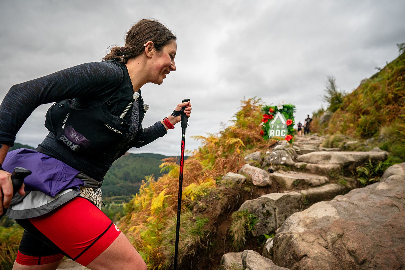A person in red shorts and a black top is hiking up a rocky trail with trekking poles. Golden ferns line the path, and a green and red marker labeled "ROC" is visible ahead. The sky is overcast, and distant hills can be seen.